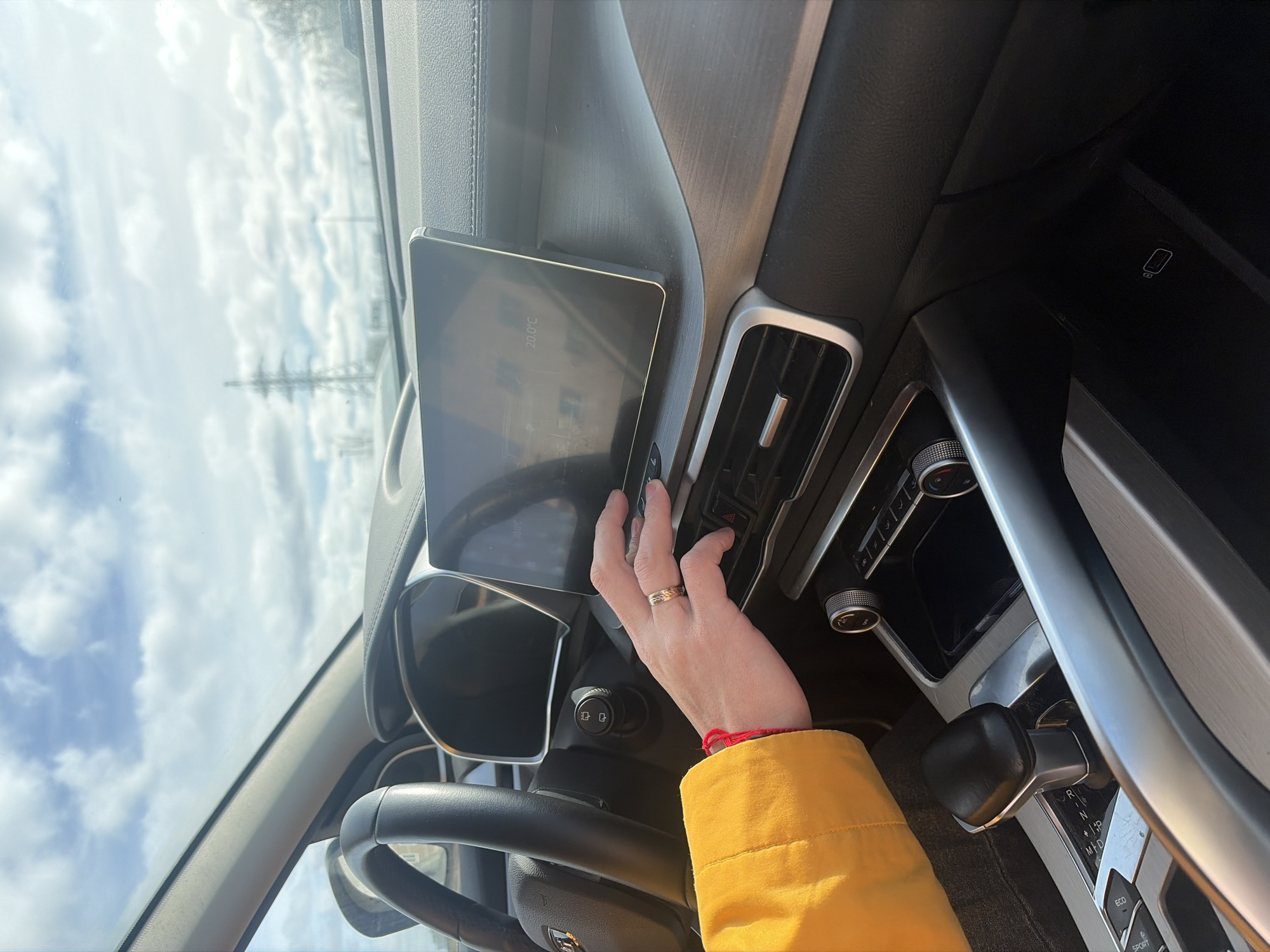 A view from inside a car shows a person's hand with a wedding ring on the ring finger reaching toward the dashboard. The dashboard has a touch screen displaying "20.0 °C." Other controls and air vents are visible, as is the steering wheel. The person is wearing a yellow jacket with a red string around the wrist. Through the windshield, the sky is visible with some clouds. The location is unknown.