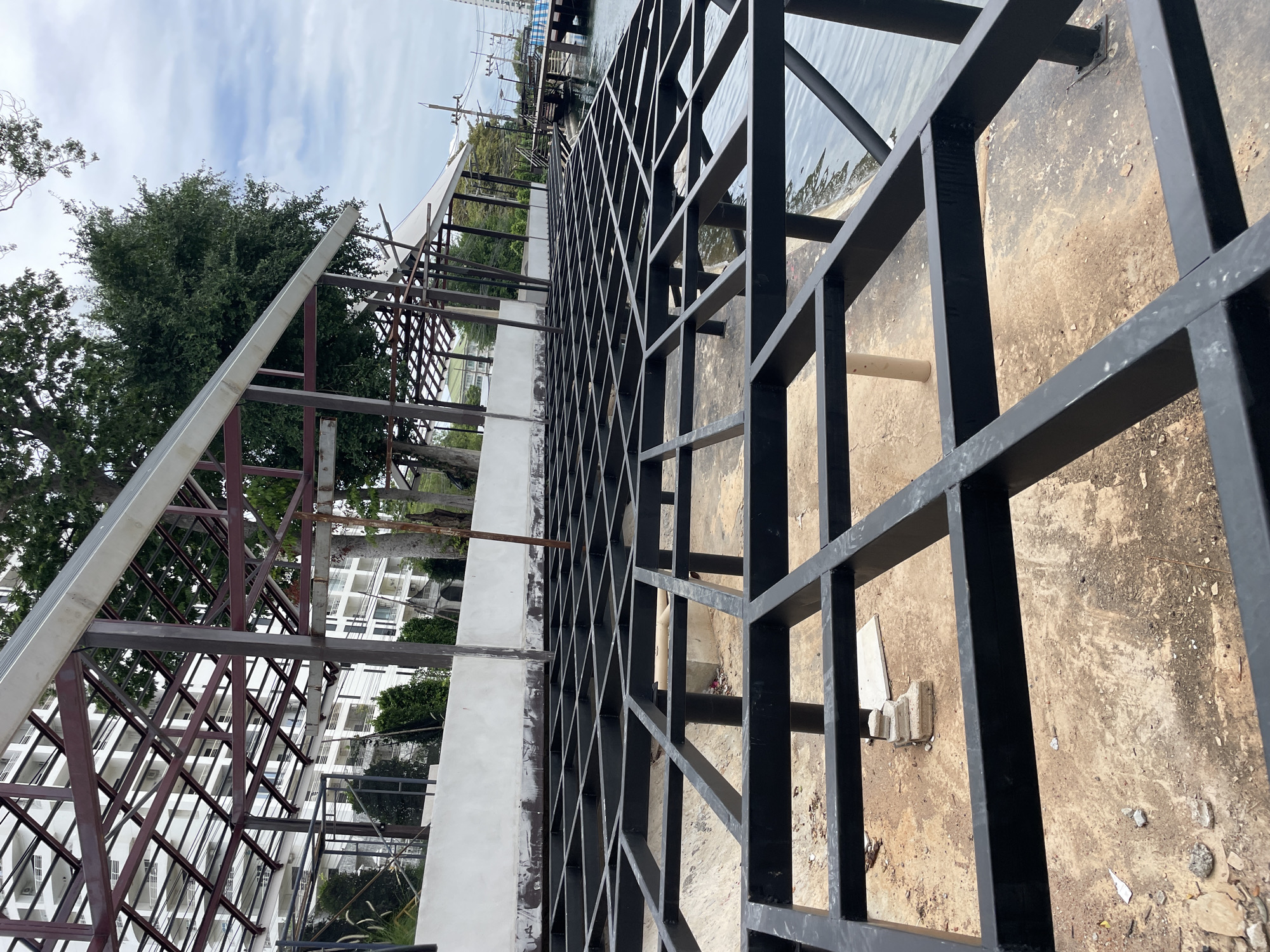 The image depicts the construction of a building or structure near the water's edge in Pattaya City, Thailand. The foreground shows a black metal grid framework, presumably for a deck or floor, laid out over a sandy and concrete base. In the background, a building with white facades and balconies is visible, along with trees and the cloudy sky. The overall scene suggests ongoing construction work, with the metal framework indicating the early stages of building. The presence of water to the right of the frame suggests a location along the coast or a canal.