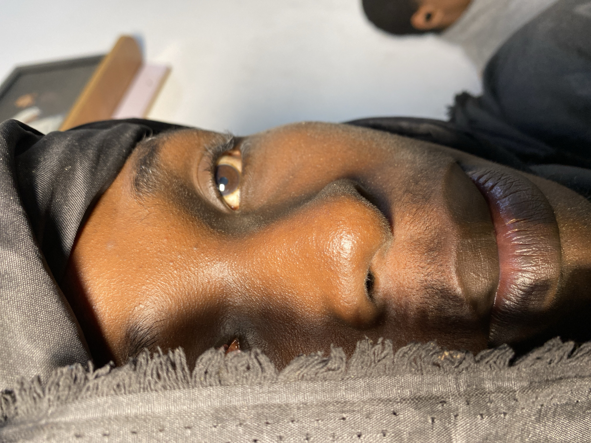 A close-up, right-facing profile shot focuses on the left side of a young Black person's face, who appears to be wearing a black head covering that drapes over their forehead and cheek. The person's brown eye is visible, with visible eyelashes. Their nose is prominent, and their lips are closed, showing texture. A dark gray scarf with a fringed edge is positioned on the left side of the frame, partially obscuring the left side of their face. The skin of the face has a warm, brown tone with subtle highlights and some visible texture.
In the background, to the right and out of focus, another person is partially visible, appearing to wear a gray hoodie and possibly an earpiece. The environment seems to be indoors, with a white wall and a wooden shelf with some items on it visible in the upper right corner. The lighting suggests an indoor setting, possibly near a window, with light casting shadows and highlighting the contours of the face. The image was captured in Bukuru, Nigeria.