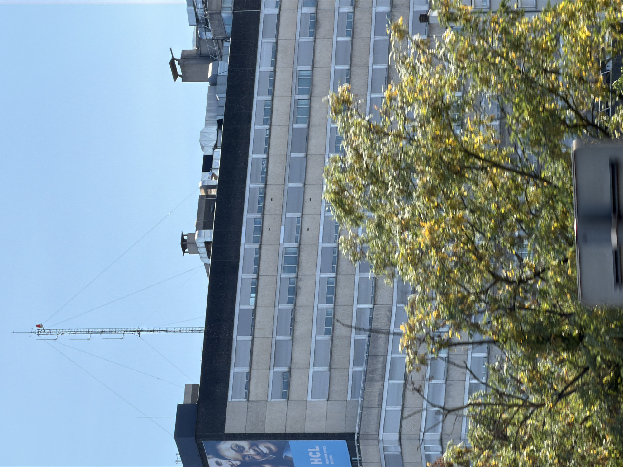 The image shows the HCL (Hospices Civils de Lyon) building in Bron, France, under a clear blue sky. The building is a multi-story concrete structure with rows of windows. A large advertisement for HCL is visible on the left side of the building. The top of the building features several ventilation systems and chimneys. A tall radio mast with antennas stands on the roof, with guy wires extending to the building. In the foreground, the blurry branches and leaves of trees with yellowing foliage suggest an autumn setting. A partial view of a vehicle is visible at the bottom of the frame.