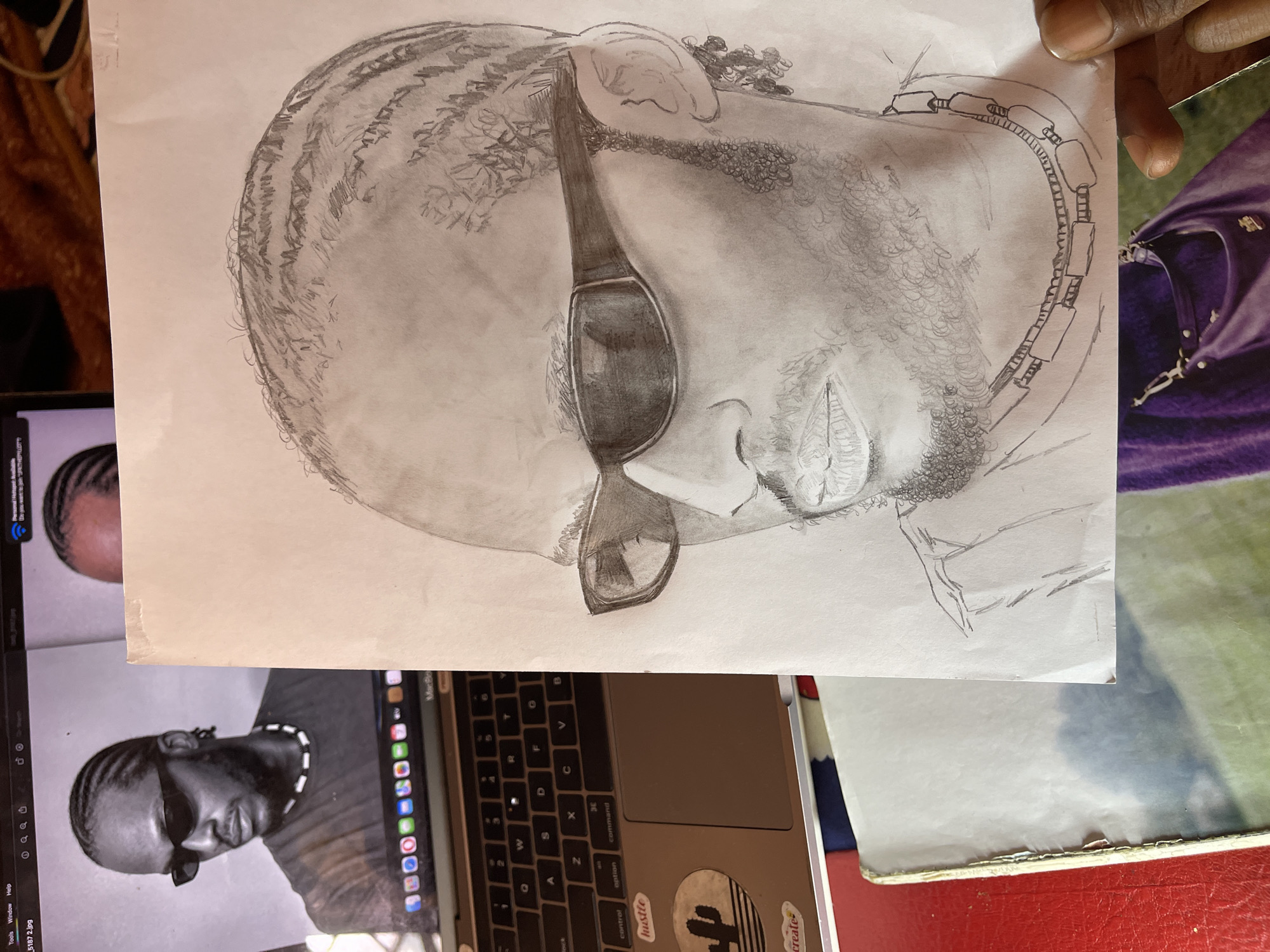 The image depicts an indoor scene in Jos, Nigeria, featuring an artistic workspace. Prominently displayed is a detailed black and white pencil drawing of a male individual, likely of African descent, rendered from the upper chest upwards. The subject wears braided hair (cornrows), dark wraparound sunglasses, and has a beard. A necklace with alternating rectangular and cylindrical beads adorns his neck. The drawing is on white paper and is partially held by a person's fingers in the lower right corner.
To the left of the drawing, an open MacBook laptop displays the black and white photograph that served as the reference for the sketch. The photograph shows the same man, also wearing sunglasses and a dark t-shirt, looking slightly to his left. The laptop screen shows an interface resembling image editing software, with menu bars and a Wi-Fi status indicator. The laptop's palm rest features several stickers, including one that reads "hustle" and another depicting a cactus silhouette. In the background, below the drawing, parts of a purple object, possibly a handbag with a metallic clasp, are visible against a green surface. A red surface is also partially visible to the lower left of the laptop.