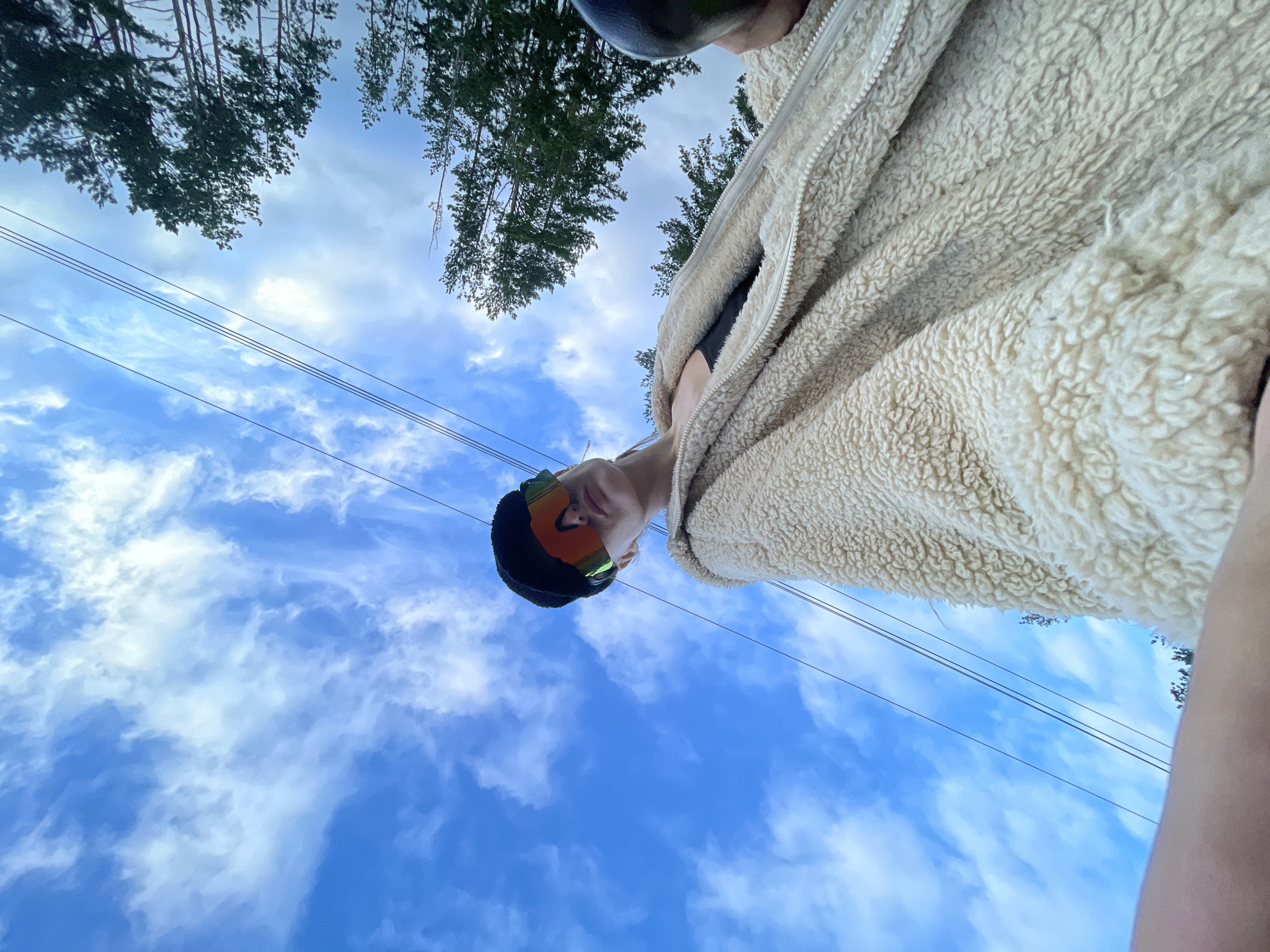In an outdoor, ground-level perspective shot taken in the vicinity of Podshivalovo, Russia, a person is viewed from below, looking up towards the sky. The individual, wearing a cream-colored fuzzy jacket over a black top and a black beanie with brightly colored ski goggles, is positioned in the foreground. The sky above is a vibrant blue, filled with scattered white clouds, and several telephone wires stretch horizontally across the frame. Trees with dark green foliage are visible at the top of the image, silhouetted against the sky. The overall impression is one of a casual moment captured outdoors.