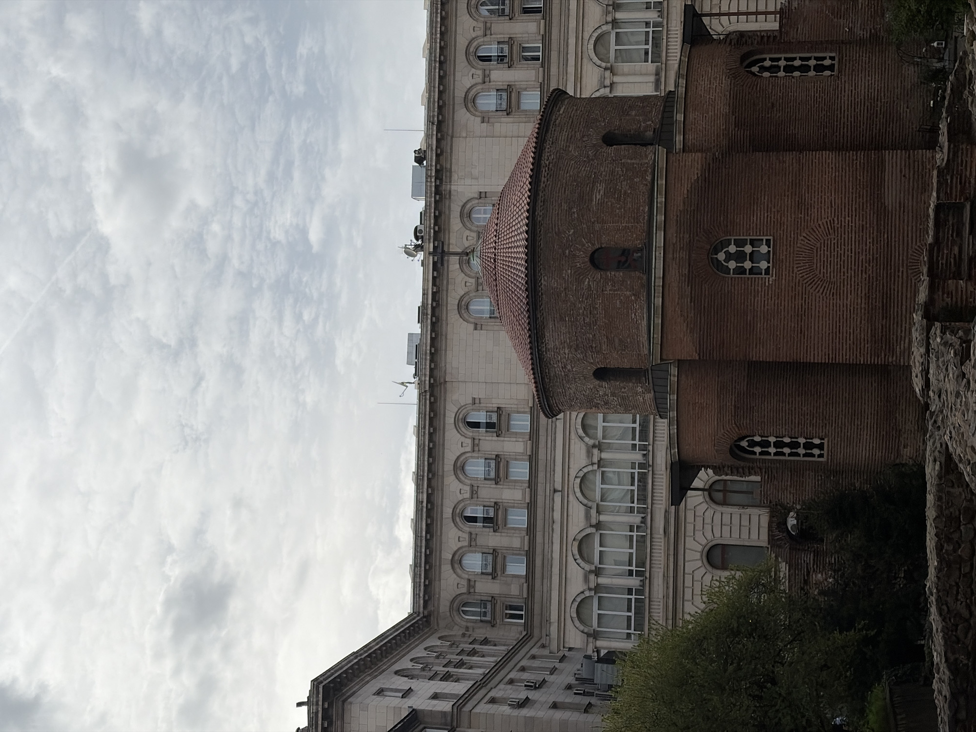 An architectural photograph presents a striking contrast between an ancient brick rotunda and a more modern, ornate stone building, under an overcast sky in Sofia, Bulgaria.
The central subject is a round, brick-built structure, possibly a church or historical monument, featuring a distinctive reddish-brown tiled dome. Its walls display intricate brickwork and include small, arched windows, some fitted with decorative grates. This style is highly suggestive of the iconic Church of St. George Rotunda in Sofia, known for its deep historical roots.
Behind and to the left of the brick rotunda stands a large, multi-story building constructed from light-colored stone. This structure is characterized by numerous arched windows, with some upper-floor windows featuring small, ornate balconies with metal railings. Various modern elements such as antennas and air conditioning units are visible on its rooftop.
The sky above is filled with soft, gray clouds, indicating an overcast day. Faint contrails are discernible in the upper left portion of the sky. In the immediate foreground on the right, a rough stone wall, likely part of ancient ruins, is partially visible, accompanied by green foliage from nearby trees or shrubs. The lighting is diffused, suggesting a day without direct sunlight. No discernible text is visible in the image.