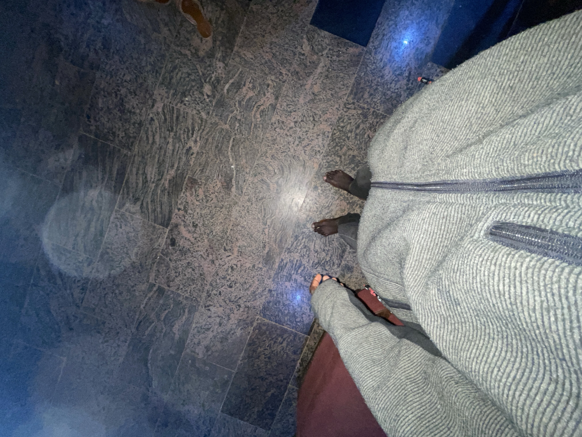 The image shows a person wearing a gray zipped jacket standing on a mottled gray and beige tiled floor. Part of another person's foot wearing a brown sandal is visible in the top left corner. The lighting appears dim with a light source creating a bright spot on the floor. A staircase or platform of dark material is partially visible in the background. Location information is not clear.