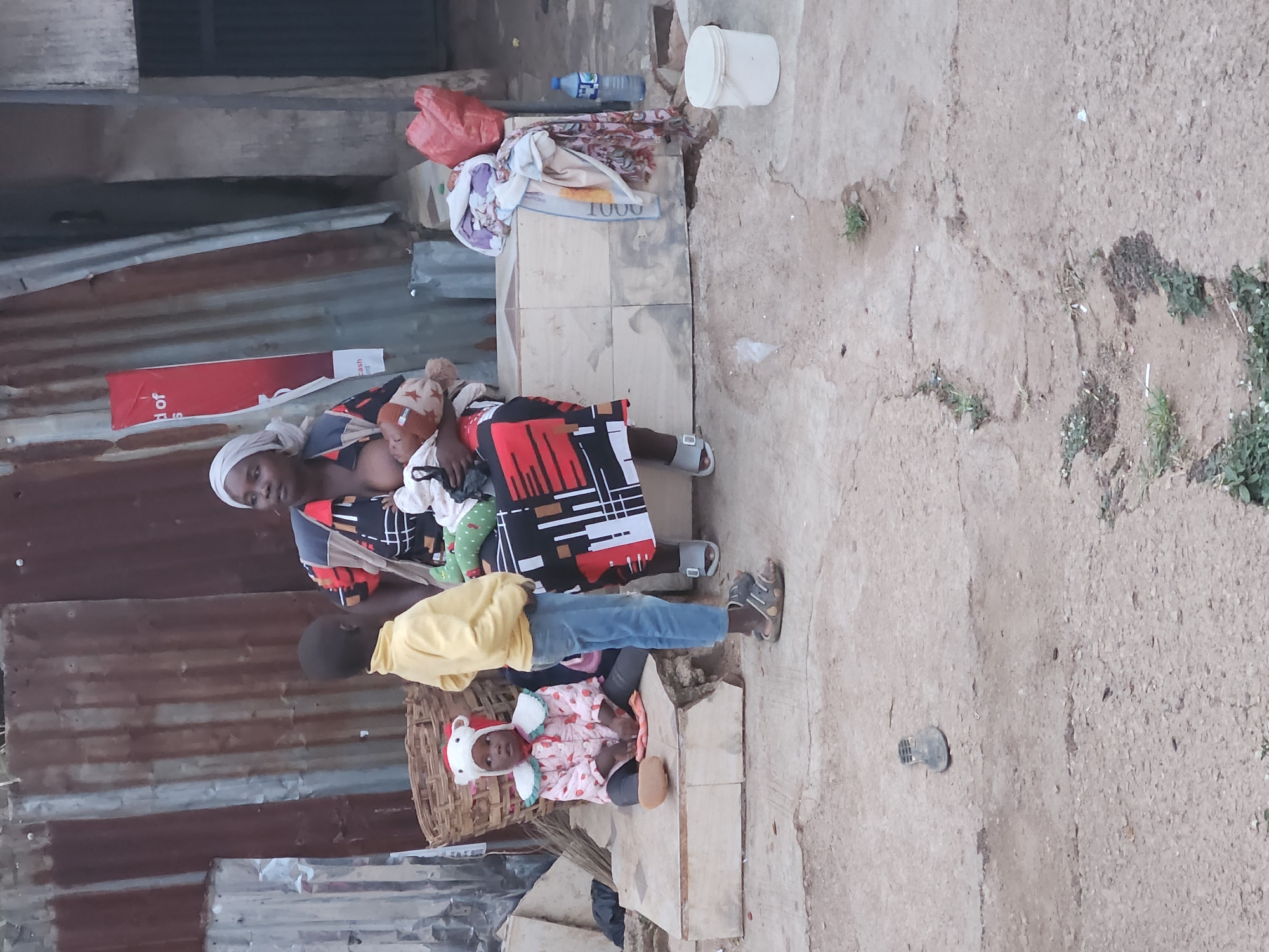 A woman, likely a mother, is seated outside a corrugated iron structure, breastfeeding an infant. A slightly older child stands beside her while a younger child, wearing a hat with bunny ears, sits on a low step or basket nearby. Various personal items are stacked behind the woman on a ledge. The ground is a mix of concrete and exposed dirt with some vegetation. A water bottle and a white bucket are also visible on the ledge. This scene takes place in Jos, Nigeria.
