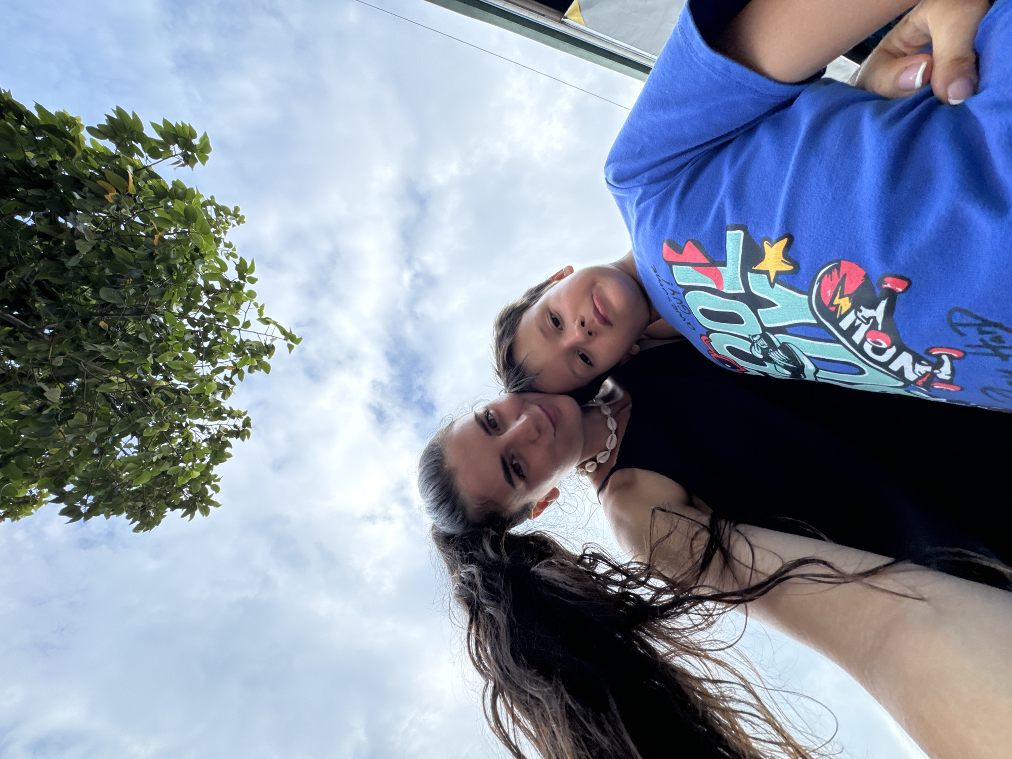 This image is captured from a very low angle, looking upwards towards the sky and subjects, with the frame rotated counter-clockwise.
Two individuals are featured prominently. In the foreground, positioned in the lower-middle and right of the frame, is an adult female. She has long, dark, wavy hair pulled back into a ponytail and is wearing a dark top. A white or light-colored shell necklace is visible around her neck. She is looking directly at the camera with a slight smile. Her arm and hand, showing neatly manicured fingernails with white tips, are visible towards the upper-right.
Slightly behind and above her, positioned in the upper-right quadrant, is a child, likely a boy, with light brown hair. He is wearing a blue t-shirt with a colorful graphic design that includes stylized text and imagery, such as a skateboard and a yellow star. Partial words like "AWESOME GUY" are discernible on the shirt. The child is also looking towards the camera with a neutral to curious expression.
The background is dominated by a bright, cloudy sky, indicating daytime. In the upper-left portion of the image, a lush green tree with numerous leaves provides a natural contrast to the sky. A thin, dark line at the very top of the frame suggests a distant building edge or roofline. The overall scene conveys a casual, outdoor setting, possibly in a tropical or subtropical environment like Phan Thiết, Vietnam, consistent with the vibrant foliage and bright sky.