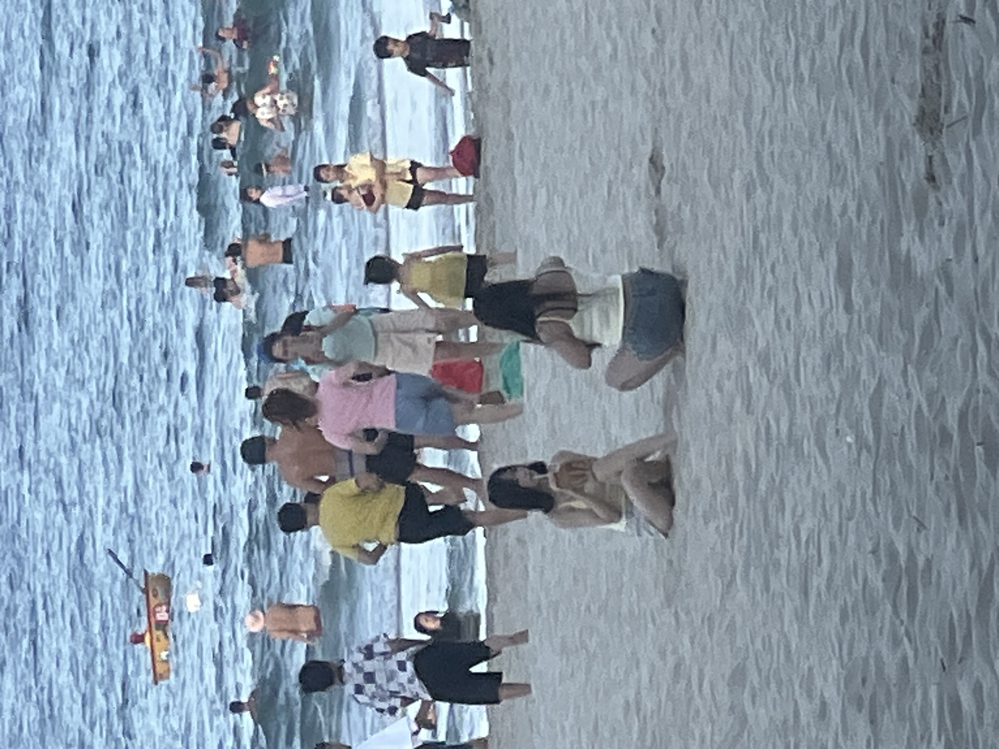 The image depicts a crowded beach scene in Da Nang, Vietnam. The primary subjects are a large group of people, many of whom are in swimwear, engaged in various beach activities. Some individuals are wading in the ocean, while others are gathered on the sand. The sea appears calm with gentle waves.
In the foreground, several people are lying or sitting on the sand. One woman in a yellow top is lying down, and another woman in a yellow top and denim shorts is sitting. Further back, a group of people in swimwear are visible, some standing and others sitting. A small boat with a red and yellow color scheme is floating in the water, with a person standing on it.
The activity taking place is a day at the beach, with people enjoying the water and sun. The scene suggests a warm, possibly sunny day, though the lighting is somewhat diffuse, hinting at it possibly being overcast or late afternoon. The overall atmosphere appears relaxed and recreational.
Notable details include the diverse group of people, suggesting a mix of locals and possibly tourists. The presence of the boat in the water is a prominent detail, as is the clear division between the sandy beach and the sea. There is no visible text in the image. The image is oriented vertically, rotated 90 degrees clockwise.