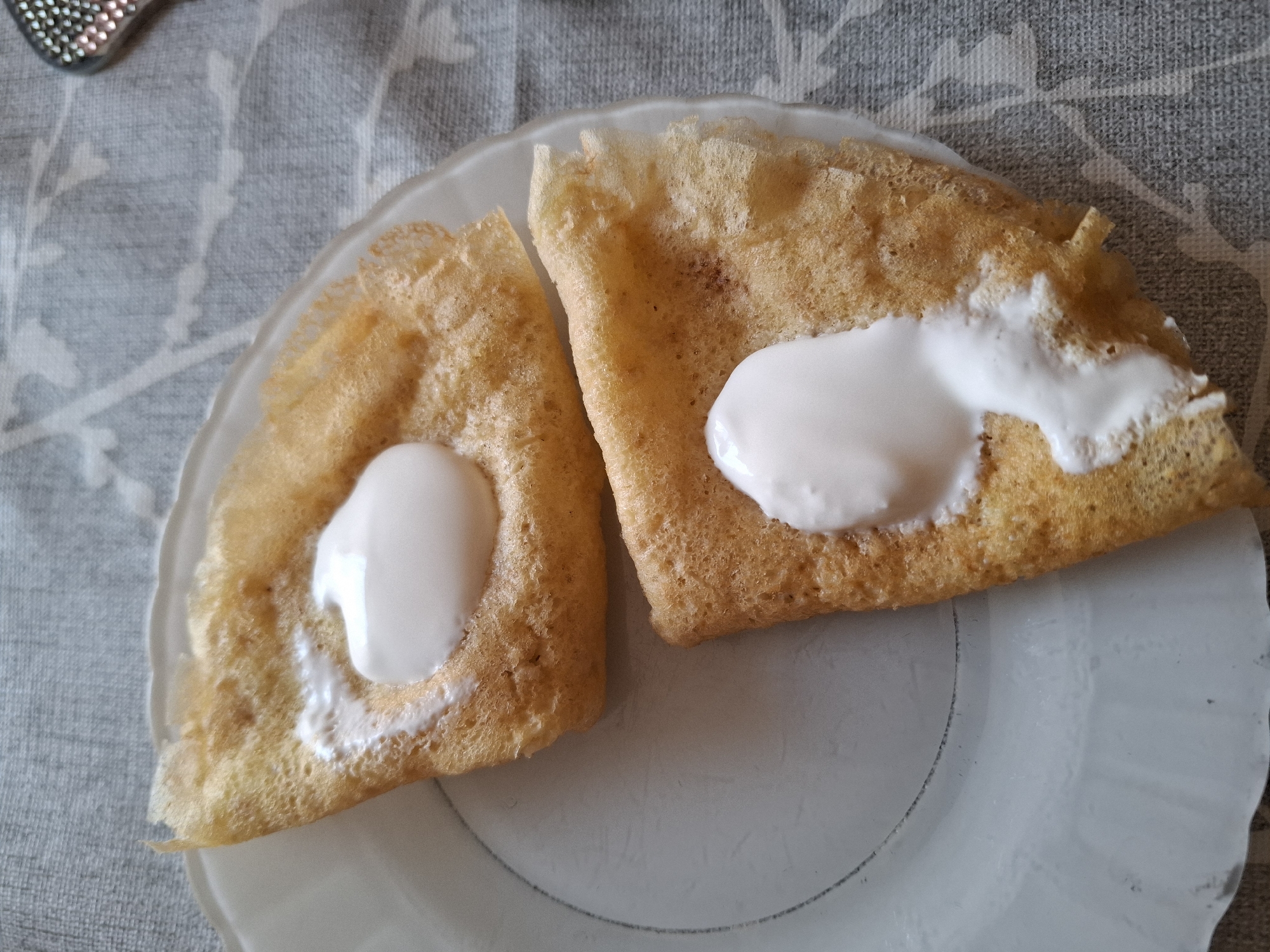 Two folded crepes with dollops of white cream are arranged on a white plate. The plate rests on a grey and white patterned tablecloth. The scene is an indoor, domestic setting. No specific location in Korenovsk, Russia can be determined from the image.