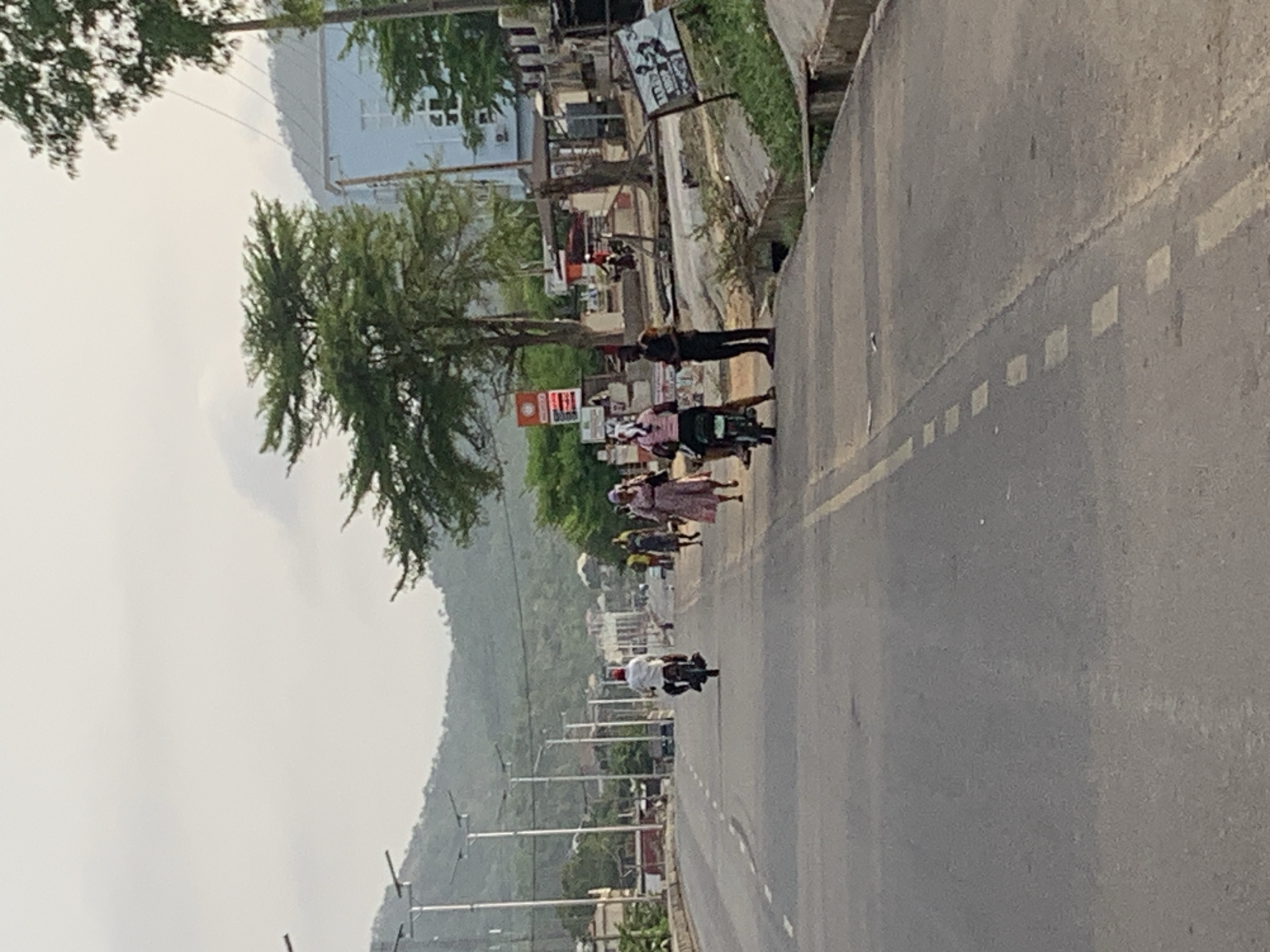 The scene depicts a roadway in Adankolo, Nigeria. The perspective is oblique, showing a street stretching into the distance with a mountain visible in the background under a cloudy sky. A few people are seen walking on the side of the road, and a motorcyclist is moving along the street. Several buildings and trees line the roadway, along with street lights. There is a white dashed line on the road.