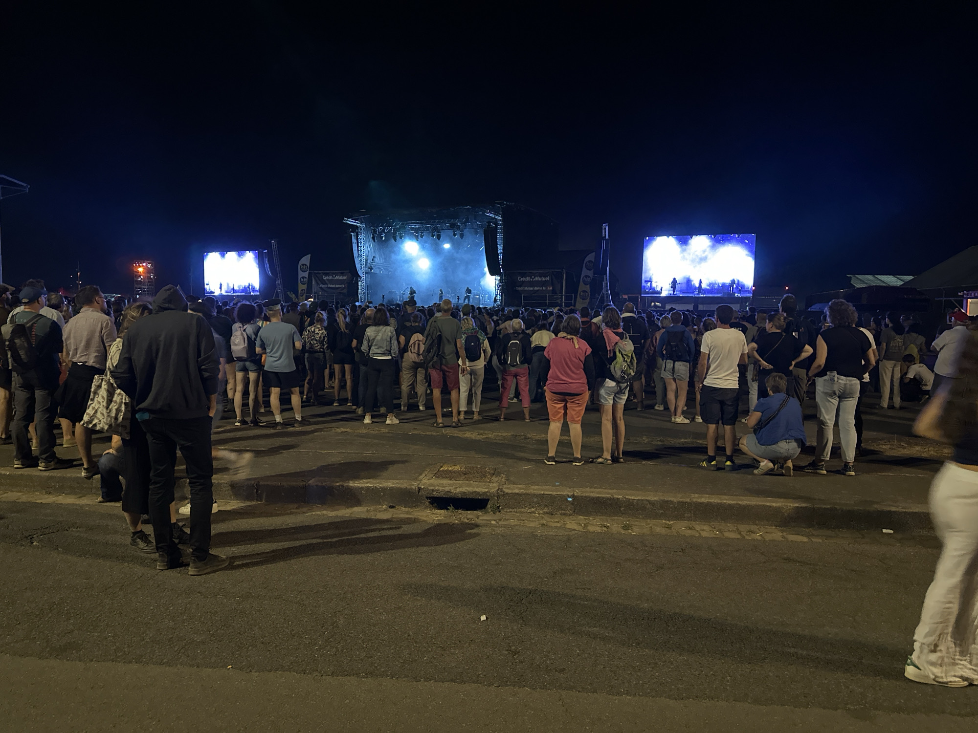 An outdoor concert is taking place at night. A large crowd of people are standing and watching a stage with bright lights. Two large screens on either side of the stage display images of the performers. The concert appears to be taking place at the Hippodrome de Pornichet, located in Saint-Nazaire, France. The ground in front of the crowd is asphalt, with a sidewalk and a drain cover visible. The overall atmosphere is lively and festive.