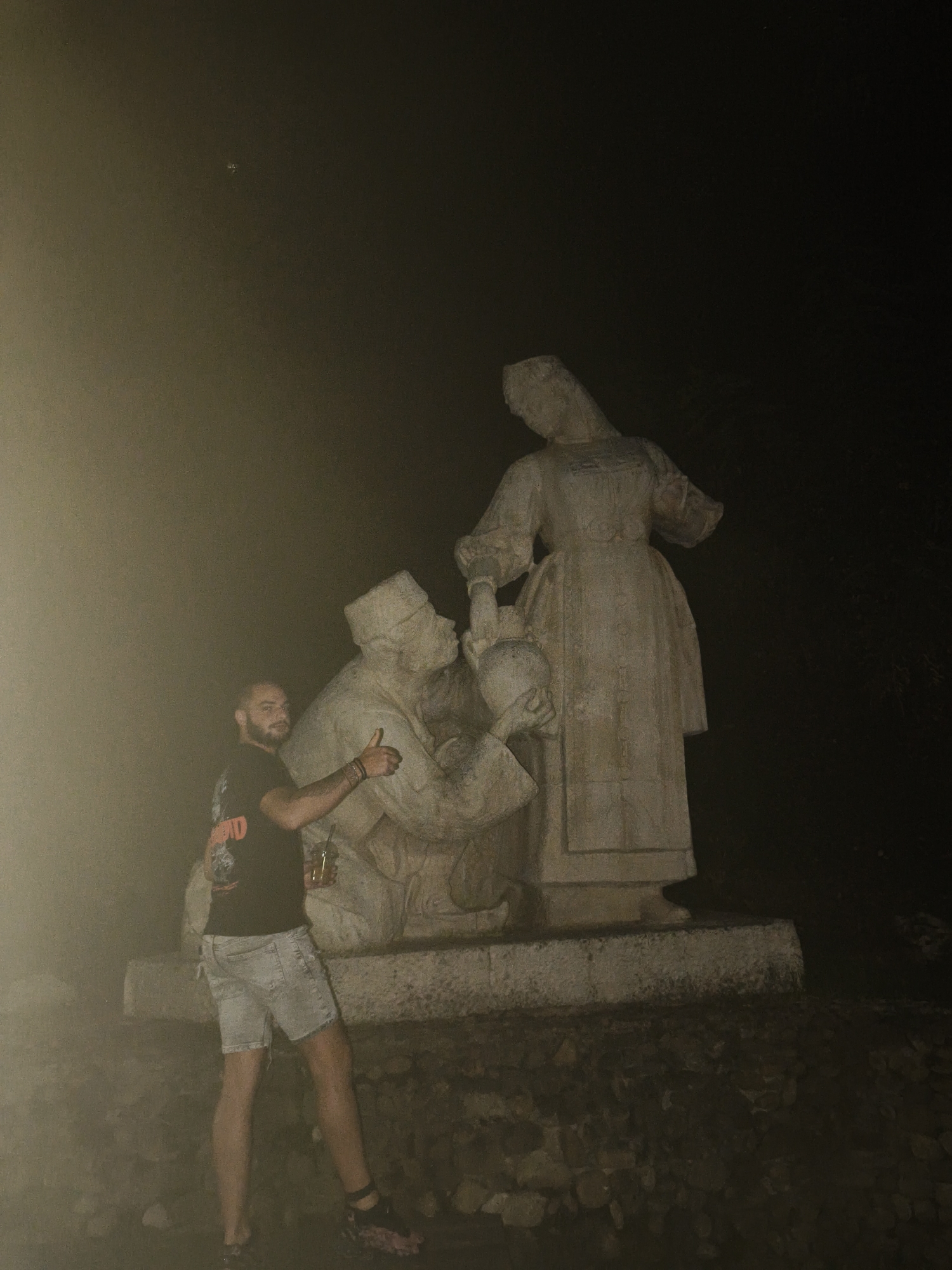 A man stands in front of a stone sculpture in Lovech, Bulgaria. The man is wearing a black t-shirt and denim shorts, and he is holding a drink in his right hand and giving a thumbs-up with his left. The sculpture depicts two figures, one kneeling and offering a jug to another standing figure, who is holding a pot. The sculpture is illuminated, and the background is dark and appears to be outdoors at night. The scene is slightly obscured by a hazy effect, possibly from fog or smoke.
