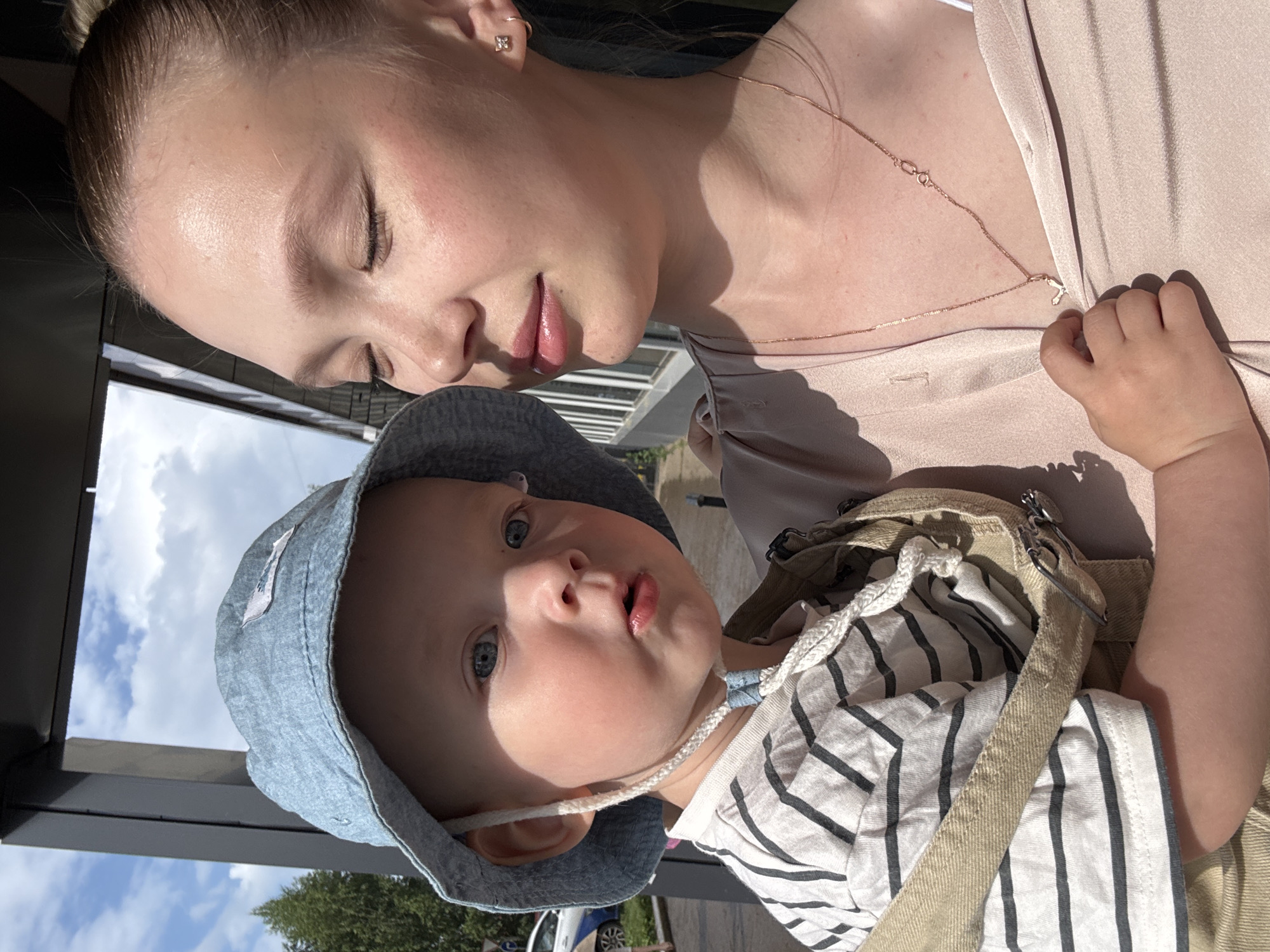 The image depicts a close-up, outdoor portrait of a young woman and a baby. The woman, with her eyes closed and a gentle expression, wears a light pink, silky blouse and a delicate gold necklace with a small charm. She has light skin and her hair is pulled back. The baby, held by the woman, wears a blue bucket hat and a striped t-shirt. The baby has blue eyes and is looking slightly to the side with an open mouth. The background shows a modern building with a dark frame and windows, and a bright blue sky with some clouds, suggesting a daytime setting. A hint of greenery and what appears to be a road with a vehicle can be seen in the blurred background to the left, indicating an urban or semi-urban environment. The lighting suggests it is a sunny day, casting shadows. The scene appears to be in Chaikovsky, Russia, based on the provided information.
