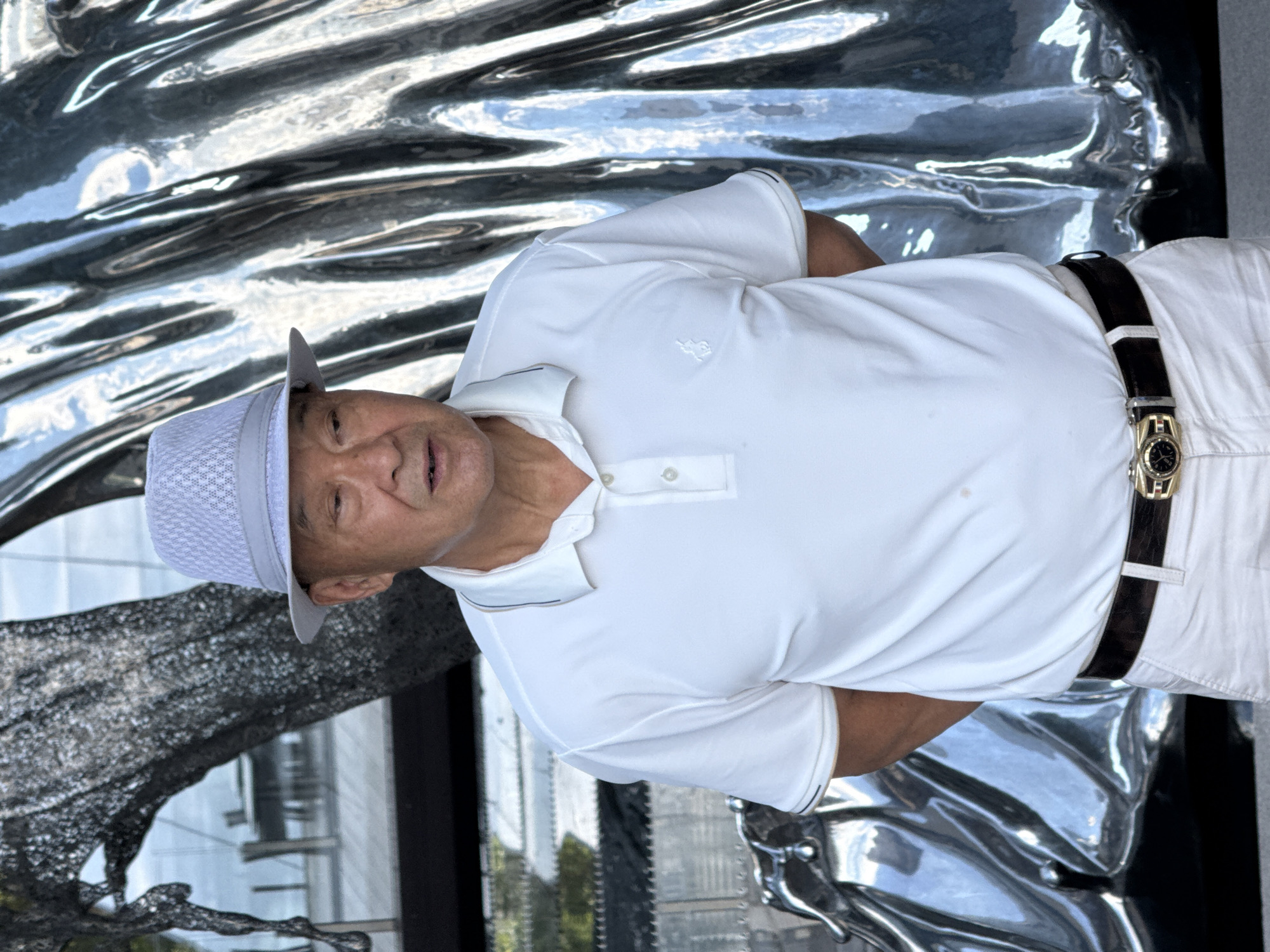A middle-aged East Asian man wearing a light blue fedora and a white polo shirt stands in front of a large, abstract, metallic sculpture. He has his hands clasped behind his back. The sculpture appears to be made of reflective, rippled material, casting a silver sheen. In the background, a blurred view of a body of water and some outdoor seating furniture suggests an outdoor setting, possibly near a waterfront. The man is wearing white pants and a dark belt with a gold buckle. The scene is well-lit, with natural light illuminating the man and the sculpture. The location is Yuzhong District, China.