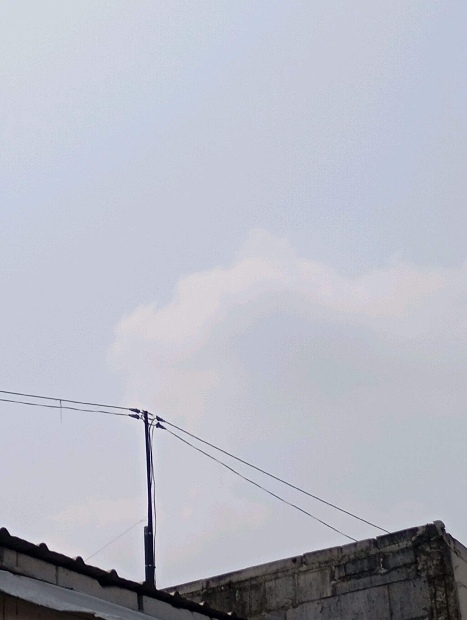 An eye-level outdoor shot shows a cloudy, overcast sky in Babelan, Indonesia. In the foreground, the top edges of two buildings are visible, one with a tiled roof and the other with a concrete block wall. A utility pole stands between the buildings, supporting overhead wires. The sky takes up the majority of the frame, with a large, wispy cloud formation visible in the upper right quadrant. The overall scene is muted and appears to be taken during the daytime, likely before or after a rain shower.