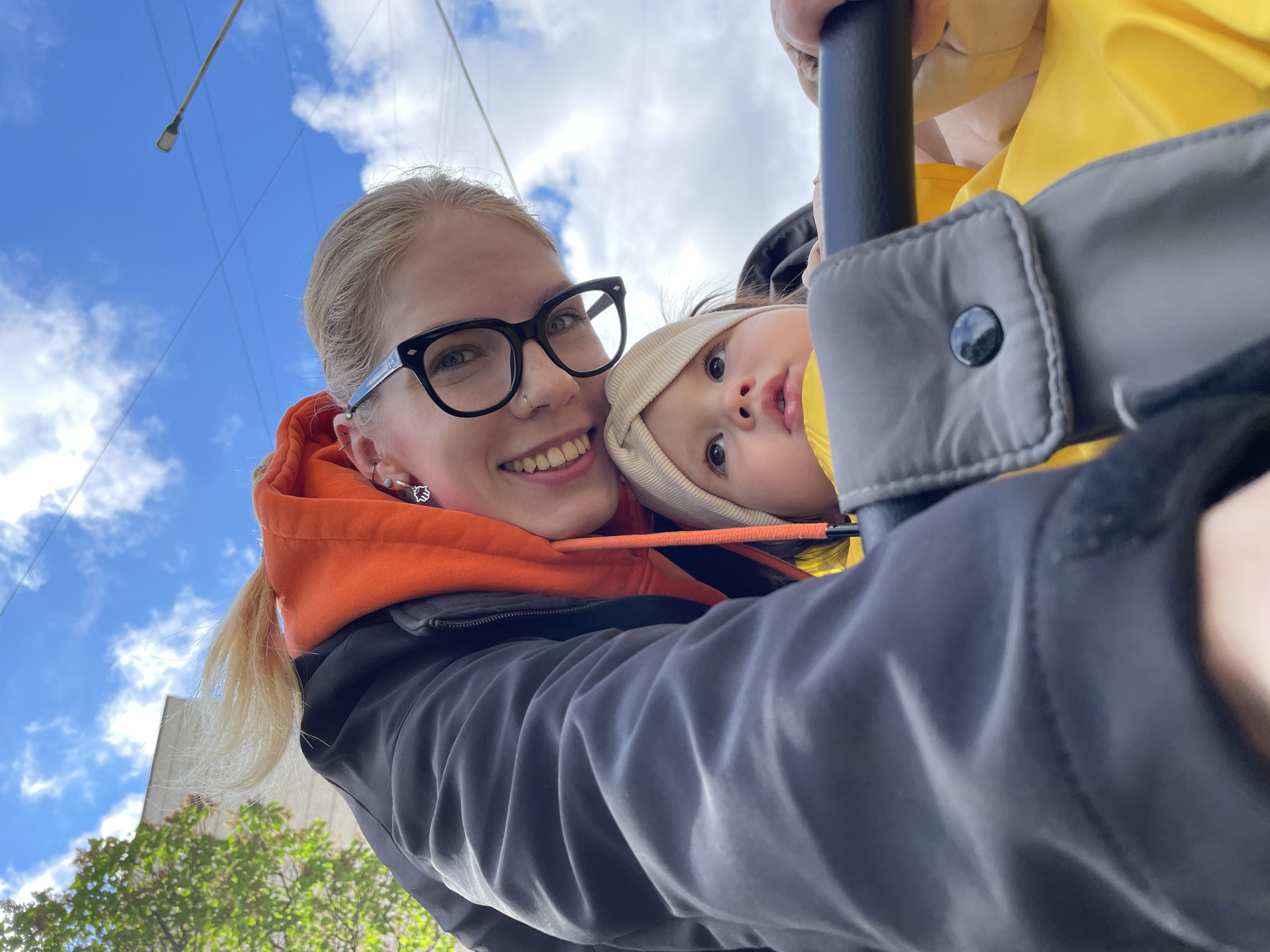 This close-up, low-angle photograph captures an adult female and a baby/toddler outdoors in Izhevsk, Russia. The adult, with blonde hair, black-rimmed glasses, and a nose piercing, smiles broadly at the camera. She wears an orange hooded sweatshirt under a dark jacket and has earrings. To her right, a baby or toddler looks upward with wide, curious eyes, partially visible from within what appears to be a stroller or carrier. The baby wears a light-colored beanie and a yellow garment.
The activity suggests a casual outing, possibly a walk, with the adult taking a selfie while interacting with the child. A dark gray padded handle or bar of a stroller/carrier is prominent in the right foreground, with a visible snap button, indicating the child is securely positioned.
Notable details include a bright blue sky filled with white clouds, suggesting a pleasant daytime setting. In the upper left background, a light pole or streetlamp and overhead wires are visible, along with glimpses of green tree foliage at the bottom and a building's edge. Both individuals appear content; the adult is smiling, and the baby's expression is one of engaged curiosity. There is no visible text in the image.