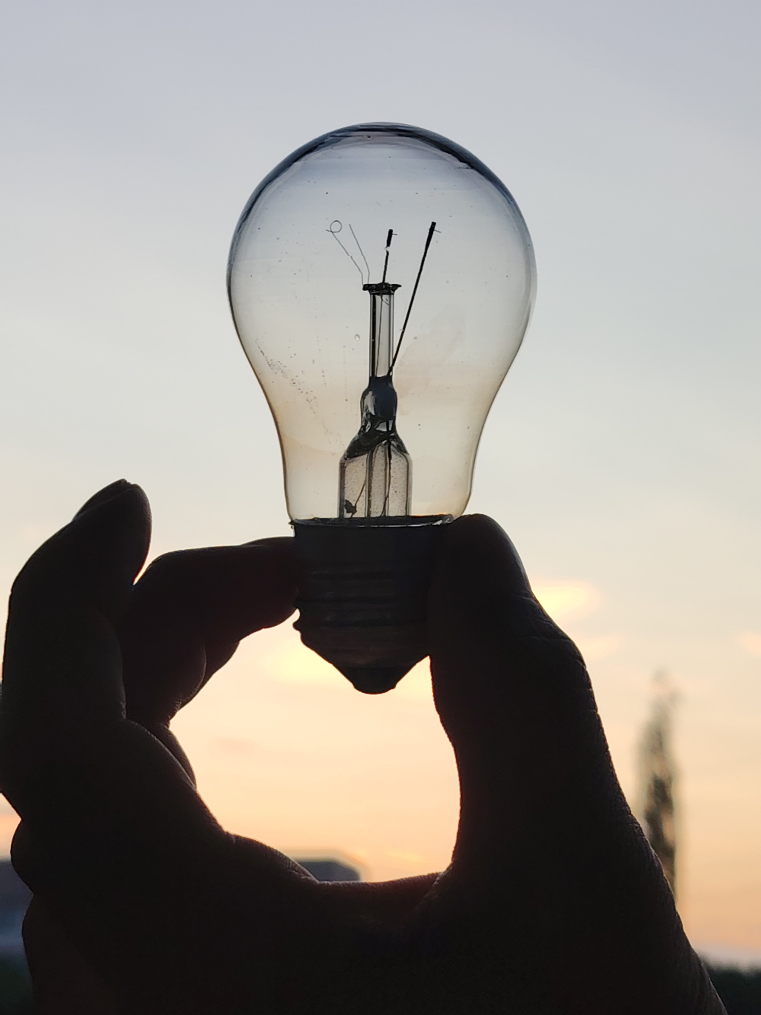 A close-up, silhouette shot captures a hand holding a clear glass light bulb against a dusky sky. The hand, silhouetted in black, cradles the base of the light bulb. The light bulb itself, though unlit, allows the faint colors of the sunset to filter through its glass. The sky behind is a gradient of soft grey and pale orange. In the far background, a blurry, dark shape suggestive of a building or tower can be discerned. This scene is observed from the city of Chaikovsky, Russia.