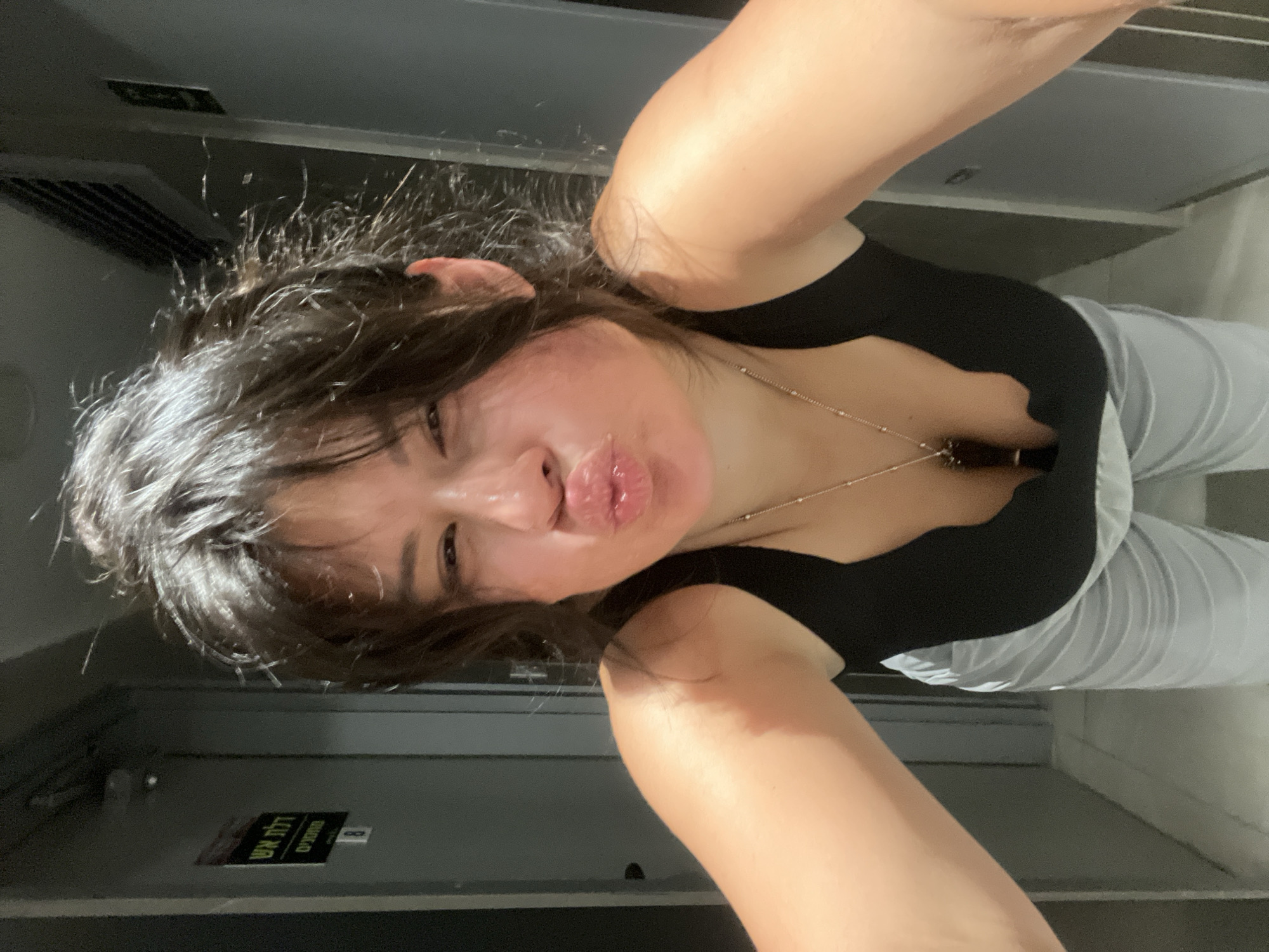 A young woman with dark hair pulled up and bangs framing her face, and light-colored pants and a black top, poses for a selfie in what appears to be a narrow hallway. Her arms are outstretched, holding what is assumed to be the phone. She has her lips pursed in a kissing gesture, and one eye is slightly squinted. A silver necklace with a dark pendant rests against her chest. In the background, to the left of the frame, a sign with Hebrew text is visible on a gray door. The hallway itself is also gray, with a tiled floor and lighting fixtures visible on the ceiling. The overall environment suggests an indoor public space, potentially a corridor in a building. The presence of Hebrew text on the sign indicates the location is in Israel, and more specifically, the text on the sign appears to be "כללית" (Klalit), which is a major health maintenance organization in Israel, suggesting the photo might have been taken in a clinic or hospital setting in Kiryat Bialik, Israel.