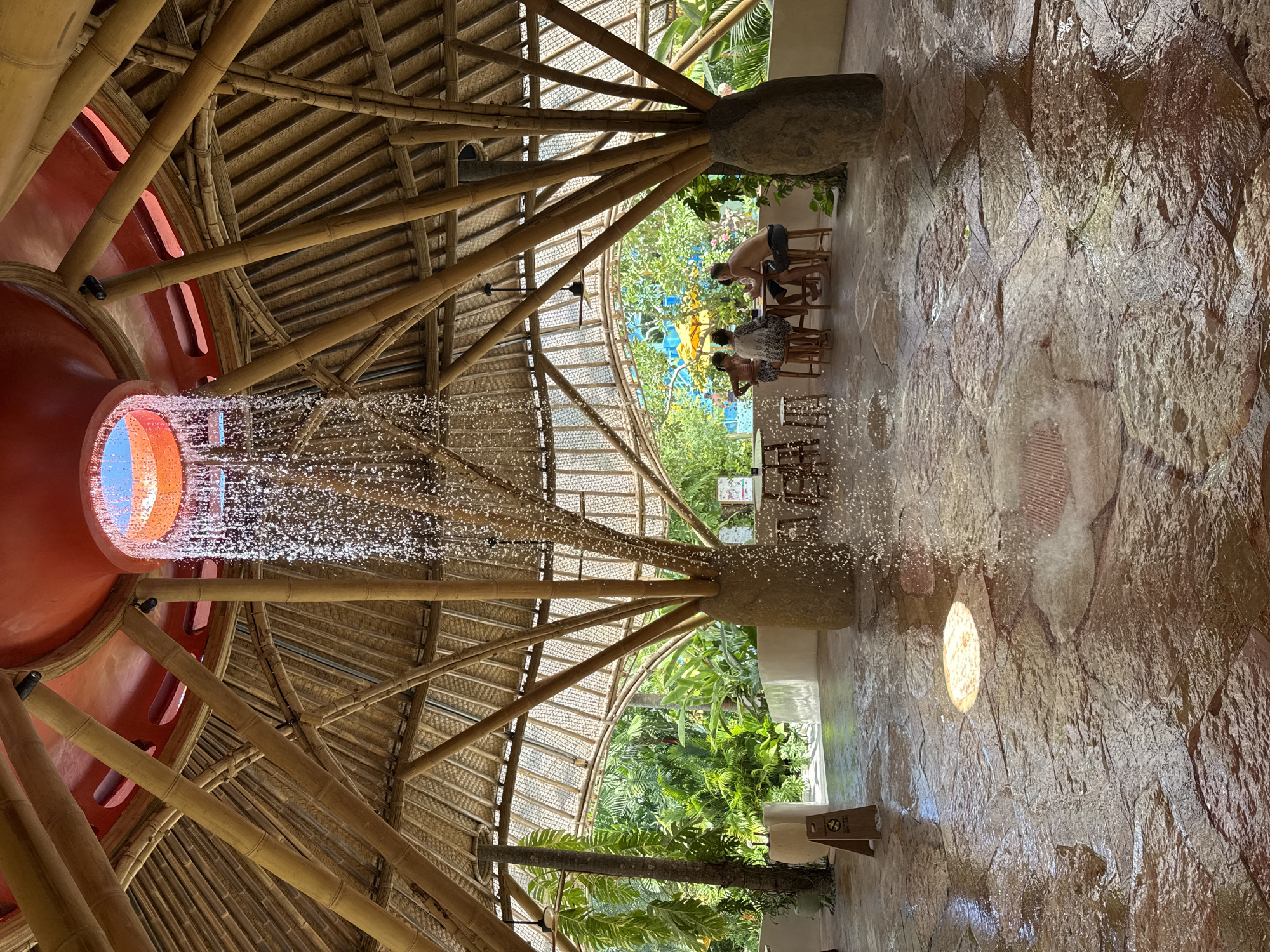 The image captures the unique architectural design of the Potato Head Studios in Kuta, Indonesia. Looking upwards, a bamboo structure supports a red, circular element with an opening that reveals the sky. Water cascades from the opening, resembling a waterfall effect. Beneath the structure, on the opposite side, a stone wall is visible. Several people are seated at tables, engaged in conversations. The surrounding environment includes lush greenery and other architectural elements, creating a blend of natural and man-made elements.