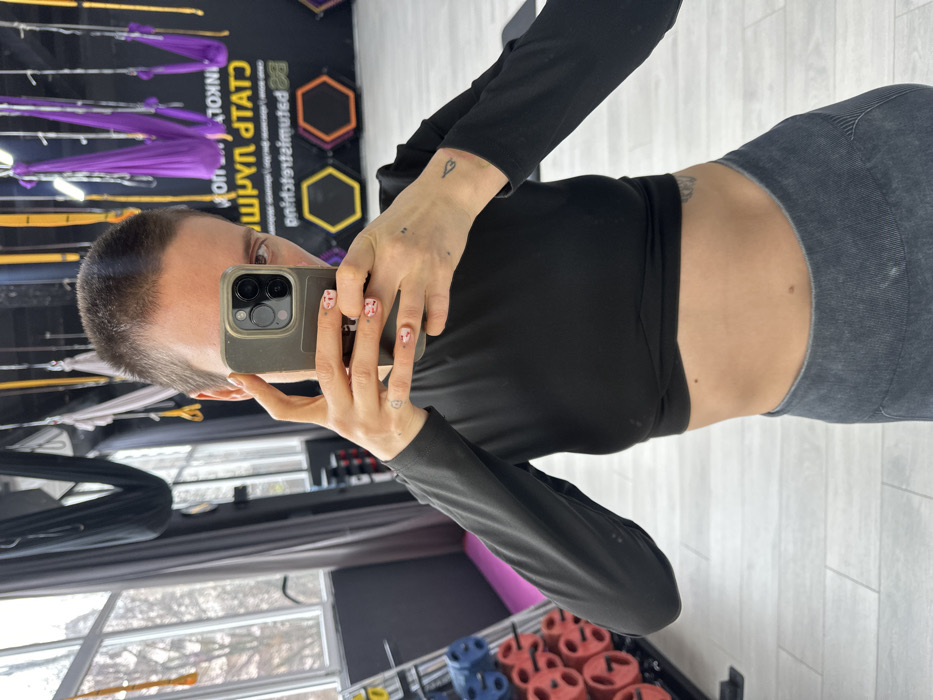 An adult individual with short, buzz-cut hair is shown from the mid-torso up, taking a mirror selfie inside a fitness studio. The individual wears a black long-sleeved crop top, exposing the midriff, and dark grey high-waisted athletic leggings. Fingernails are painted with a light base and reddish abstract patterns. A small heart-shaped tattoo is present on the left hand/wrist, and a smaller, dark tattoo on the right wrist. A dark-colored smartphone, likely an iPhone 15 Pro/Pro Max due to the visible camera configuration and dynamic island reflection, is held centrally, obscuring most of the face.
The environment is a gym or fitness studio in Batumi, Georgia. The rear wall to the right is black, featuring mirrored yellow text that reads "BE STATUMGYM" and "ფინქოლ სტუდიო" (Pinkol Studio), along with yellow and orange/red outlined hexagonal designs. Overhead, purple and yellow fabric hammocks, consistent with aerial fitness equipment, are suspended from the ceiling. To the left, large windows provide natural light, revealing an overcast exterior. A black rack holding various colored circular weights (red, blue, grey) is partially visible in the lower left. The floor is covered with light-toned rectangular tiles arranged in a herringbone pattern.