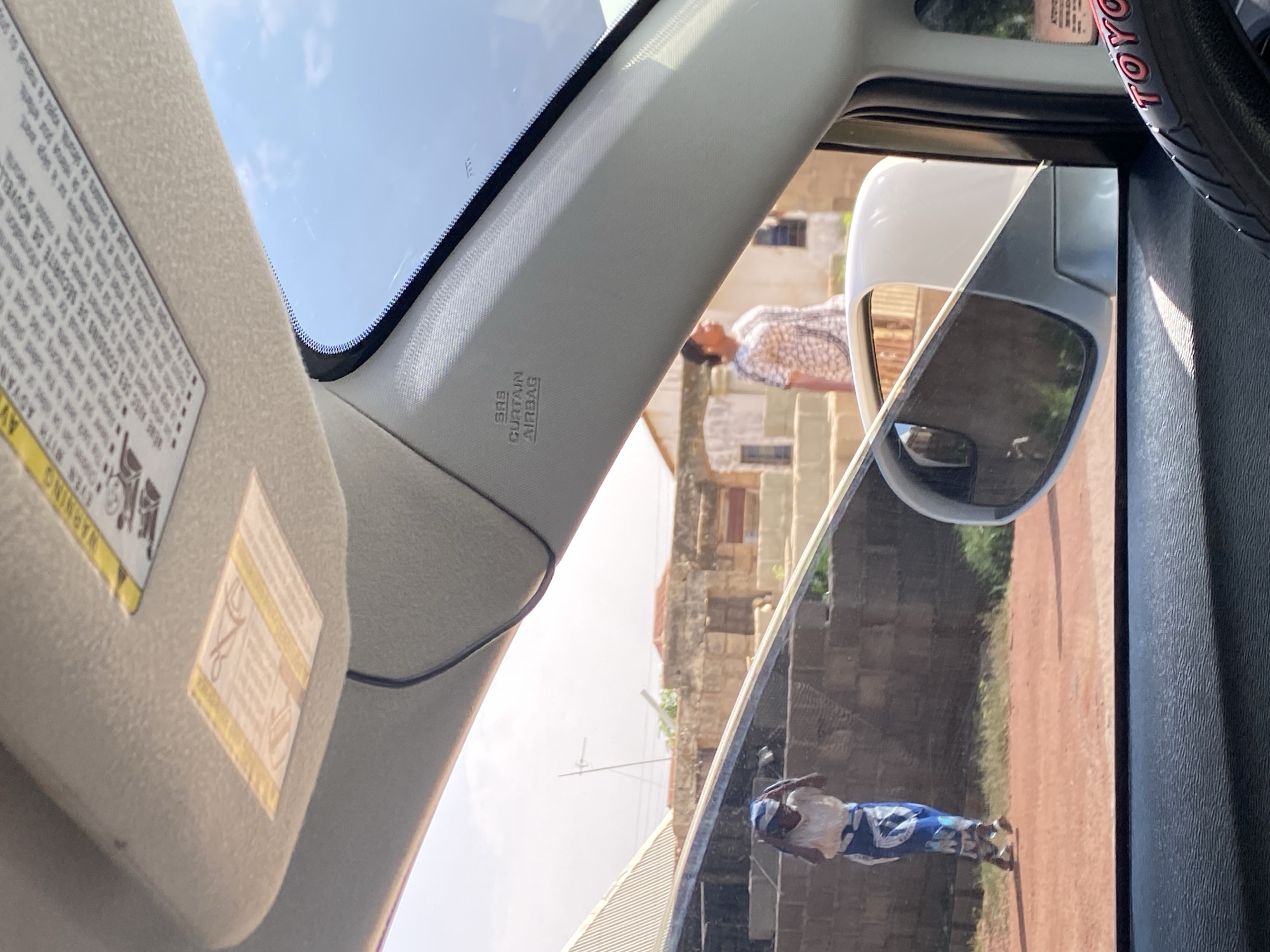The image, taken from inside a vehicle, shows a view of the city of Bar Jirgi Summa, Nigeria. The foreground is dominated by the car's interior, including the sun visor and a portion of the steering wheel with a "TOYOTA" logo. A side mirror reflects the exterior scene.
In the middle ground, the exterior environment is visible through the car window. Two women are present. One woman, on the left, is walking and appears to be adjusting her headscarf. She is wearing a white top and a blue and white patterned skirt. Behind her, a second woman, on the right, is also walking and is dressed in a patterned dress. The background consists of buildings constructed from what appears to be stone or brick, with some signs of wear and tear. The ground is reddish-brown, suggesting a dirt or unpaved road. The sky is bright blue with some clouds. A sun visor sticker with a warning symbol and text is visible on the car's interior. The "SRS CURTAIN AIRBAG" label is also visible on the car's interior pillar.