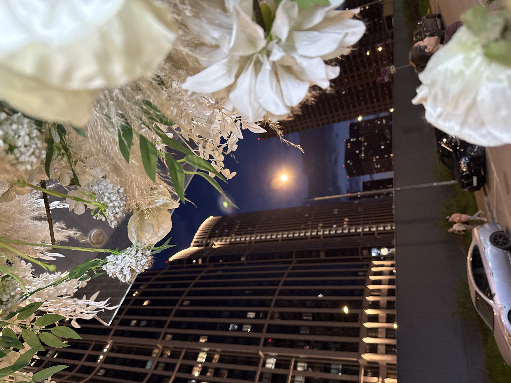 The image depicts a night scene in an urban environment, seemingly taken from a low angle, looking upwards towards tall buildings. White floral arrangements with greenery dominate the foreground, partially obscuring the view. The background features lit buildings with numerous windows, stretching into the night sky. A bright light, possibly a street lamp or the moon, is visible between the buildings. On the right side of the image, a person is seen standing beside a car, with a street and some greenery. The location is in an urban area and NOT Khimki, Russia.