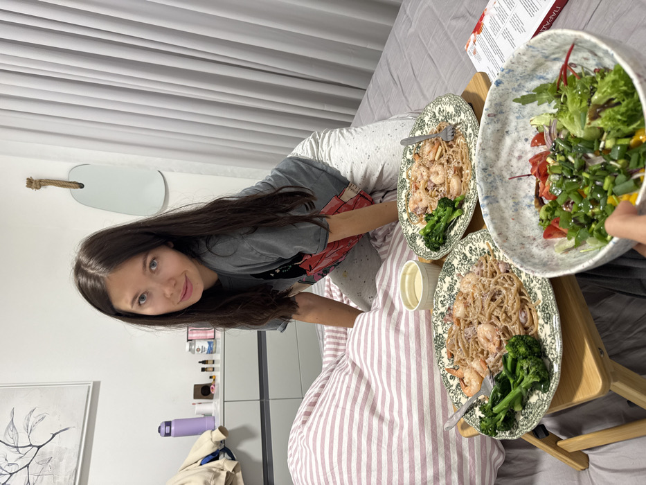 The image depicts an indoor scene, likely a bedroom, with a young adult female on a bed surrounded by food. The exact location within Tel-Aviv, Israel, is not identifiable from the visual evidence.
The subject is a female with long, dark hair, wearing a grey t-shirt with a graphic design and light grey patterned sweatpants. She is positioned on her knees on the bed, facing and looking directly at the camera with a subtle smile. Her right arm is visible, resting on the bed.
The bed is covered with light grey fitted sheets. To the left, a portion of a duvet or blanket with pink and white stripes is visible. A small, light-colored wooden bed tray is centrally placed on the plain grey sheet, holding two patterned plates. Each plate contains a serving of pasta with shrimp and broccoli, accompanied by a silver fork. Between the plates on the tray, a small white ramekin or cup holds a light-colored liquid. To the right of the tray, an open book or magazine with a white cover and visible text, including Greek characters "ΠΑΡΑΣ...", lies on the sheet. In the immediate foreground, a hand is holding a large, speckled bowl filled with a salad composed of mixed greens, chopped tomatoes, corn, and green onions, topped with dressing.
The background features a white wall. To the left, a light grey chest of drawers or nightstand is visible. On its surface, a purple insulated water bottle, a beige hooded garment, several small bottles (possibly essential oils), a small brown box, and a white bottle (possibly supplements) are arranged. Above the drawers, a framed minimalist artwork depicting a dark-colored branch with leaves hangs on the wall. To the right, an irregularly shaped mirror with a thick rope hanger is affixed to the wall. Further right, light grey vertical blinds or curtains cover a window or wall section.