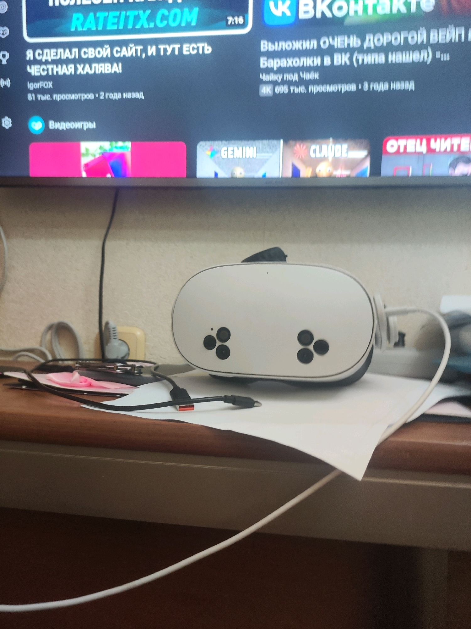 This is a photograph of a white VR headset with black buttons on a desk. The headset is connected to a charging cable. In the background, a television screen displays various videos and images, including what appears to be a VKontakte (VK) interface. One video title visible is "Я СДЕЛАЛ СВОЙ САЙТ, И ТУТ ЕСТЬ ЧЕСТНАЯ ХАЛЯВА!" which translates to "I MADE MY OWN WEBSITE, AND THERE'S HONEST FREEBIES HERE!". Another video title on the screen is "ОТЕЦ ЧИТЕ". The environment appears to be an indoor setting, likely a home or office. There are no people visible in the image. The presence of a VR headset and a television suggests a personal entertainment setup. The specific location in Kizilyurt, Russia is not discernible from the image itself.
