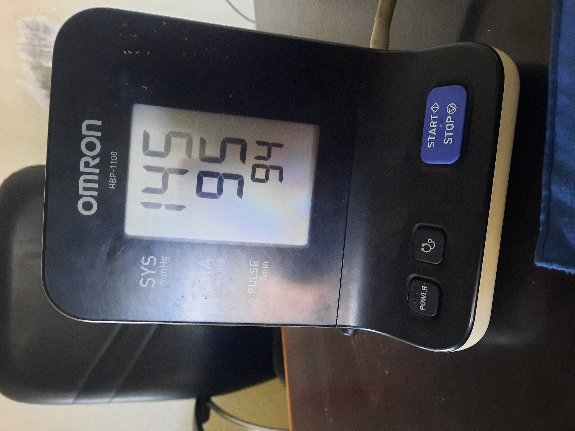The image displays a close-up, eye-level shot of an Omron HBP-1100 blood pressure monitor on a dark wooden desk. The monitor's digital screen shows a systolic blood pressure reading of 145 mmHg, a diastolic blood pressure reading of 95 mmHg, and a pulse rate of 94 beats per minute. The device is black with a beige base and features several buttons: a "POWER" button, a button with a stethoscope icon, and a "START/STOP" button. The background is blurred, showing a dark chair and a wall with some textured debris, suggesting an indoor setting. The scene is static, and no individuals are present. There is no information to suggest the location is in Lhokseumawe, Indonesia.
