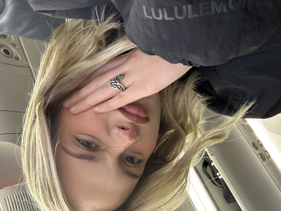 A blonde woman grimaces in an airplane seat, covering one eye with her hand. She has fair skin with freckles, blue eyes, and a nose ring. Her lips are slightly parted. She wears a large silver ring on her hand. In the background is the interior of an airplane, including a black jacket with a "LULULEMON" logo visible on the collar. The image is rotated, giving a tilted perspective.