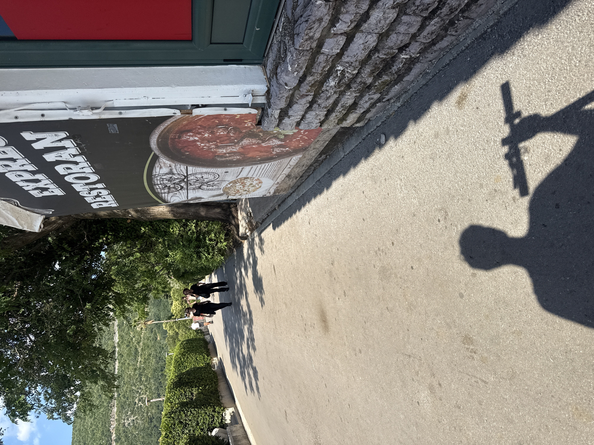 A sidewalk is seen in the foreground, with the shadow of a person holding a camera extended across it. In the middle ground, two people wearing black clothing are walking away from the camera. To the right, a sign for "EXPRESS RESTORAN" is visible, featuring an image of food. Behind the sign is a building with a green door and a red panel. The background reveals a lush green hillside with trees and a winding road. The scene appears to be in a Mediterranean coastal town, likely Budva, Montenegro, given the architectural style and vegetation.