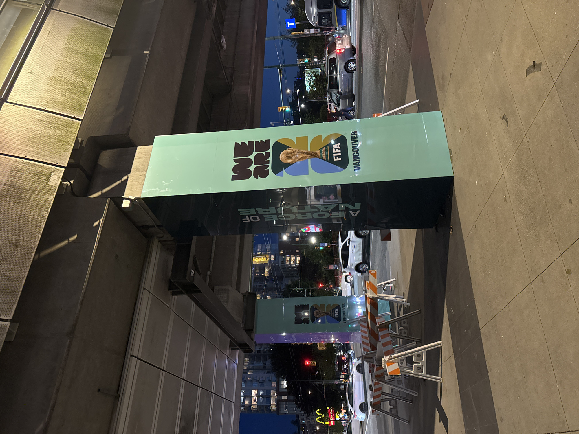 The image shows a street scene at night in Vancouver, Canada, indicated by the "VANCOUVER" text on a large, rectangular promotional sign for the FIFA World Cup. The sign is positioned along a city sidewalk, with a building on one side and the street on the other. The sign features the FIFA logo and the tagline "WE ARE A FORCE OF NATURE". The street has traffic, including cars and what appear to be construction barriers along the curb. Streetlights and building lights illuminate the scene. There are no people readily visible in the main focus of the photograph. The photo seems to be taken at ground level looking upwards. The perspective is slightly tilted.