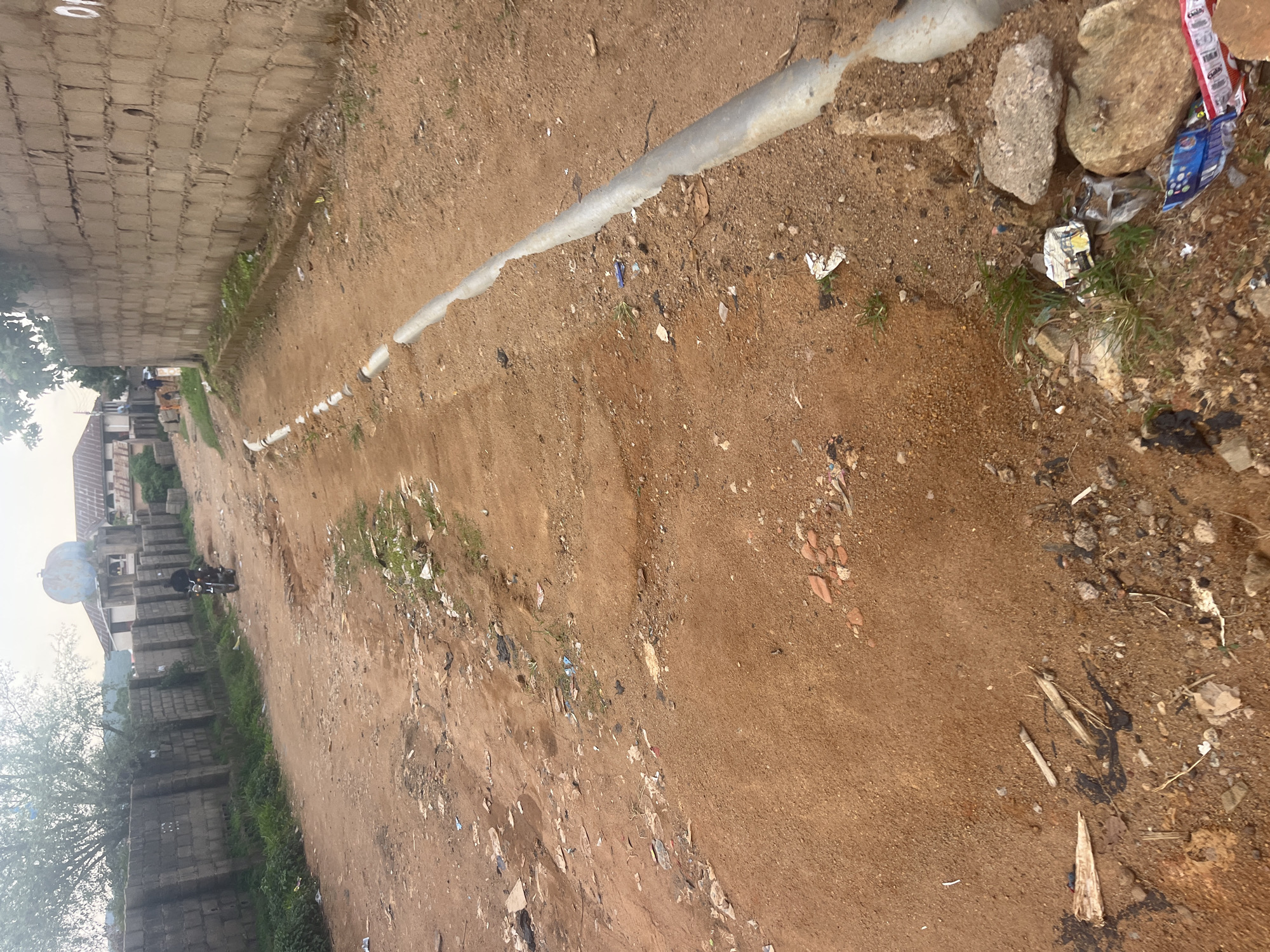 This image captures a dusty, unpaved road in Jos, Nigeria, characterized by its reddish-brown soil and scattered debris. The road is bordered on the left by a low, rough-hewn brick wall topped with overgrown vegetation. On the right, a concrete curb runs along the edge of the road, showing signs of wear and tear. Buildings are visible in the background, suggesting a semi-urban or residential area. A single motorcycle rider is seen moving away from the camera on the left side of the road, while two other individuals are walking in the distance on the right. The overall impression is one of a somewhat neglected urban environment.