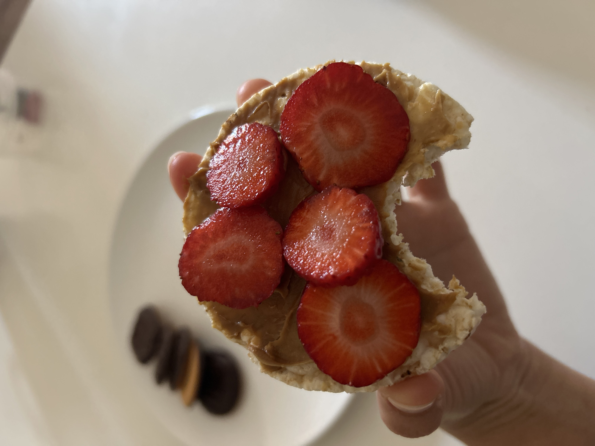A hand holds a partially eaten rice cake spread with peanut butter and topped with sliced strawberries. On a white plate in the background are several chocolate sandwich cookies. The location is not identifiable as Barcelona, Spain.