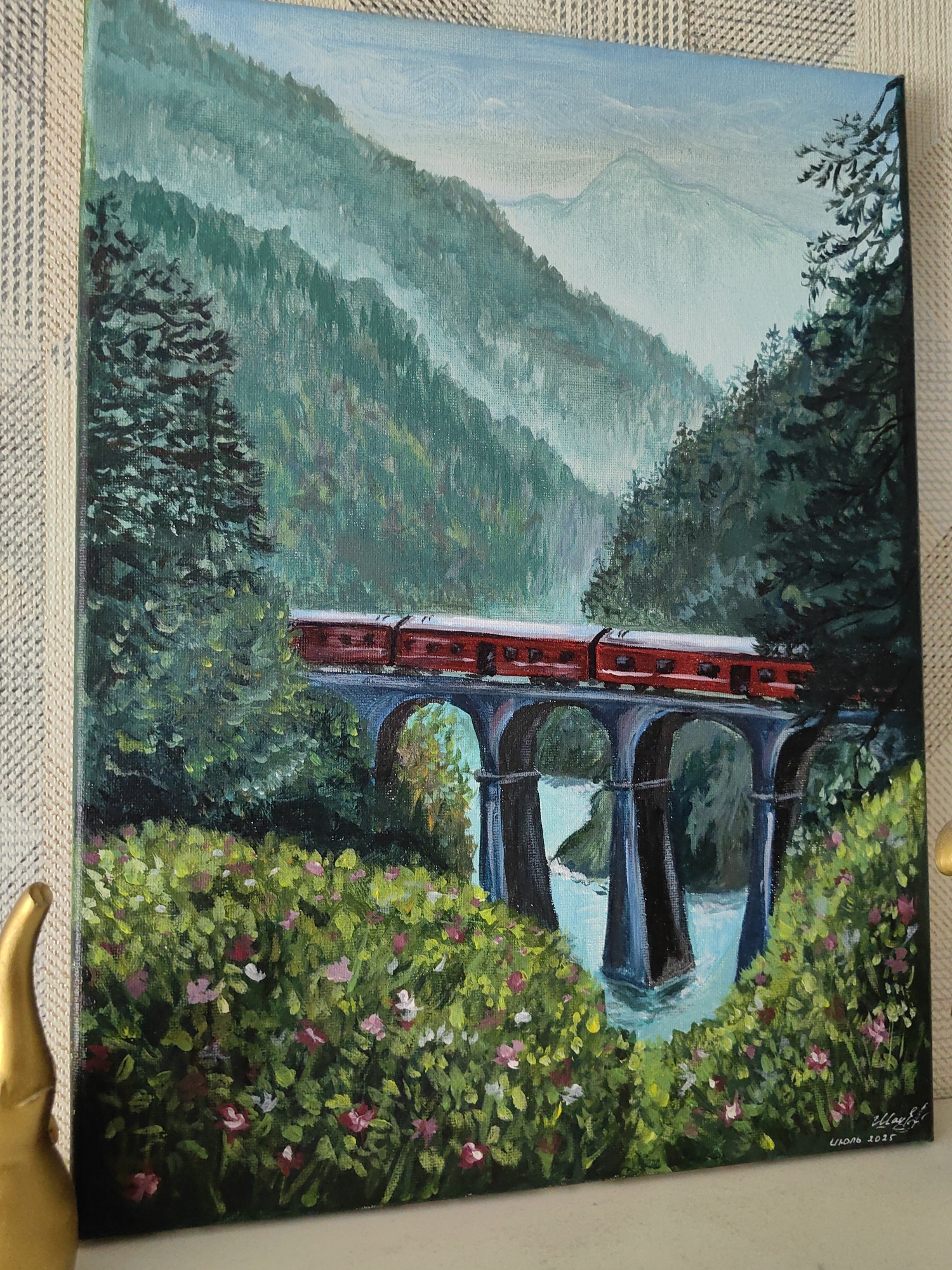 The image is a painting of a train crossing a viaduct over a river in a mountainous, forested landscape. A bright red train with multiple carriages is depicted moving from left to right across a stone viaduct with arched supports. The viaduct spans a vibrant blue river, the water of which is slightly obscured by the bridge's reflection. Lush green foliage, including trees and flowering bushes with pink and white blossoms, frames the foreground and sides of the scene. In the background, misty mountains rise, their peaks softened by atmospheric perspective. The sky above is a pale blue with subtle cloud formations. The painting is signed in the lower right corner with Cyrillic script and the year "2023". The painting is displayed against a textured beige background. The bottom left corner of the image shows a portion of a gold-colored, sculpted object, possibly a decorative item.