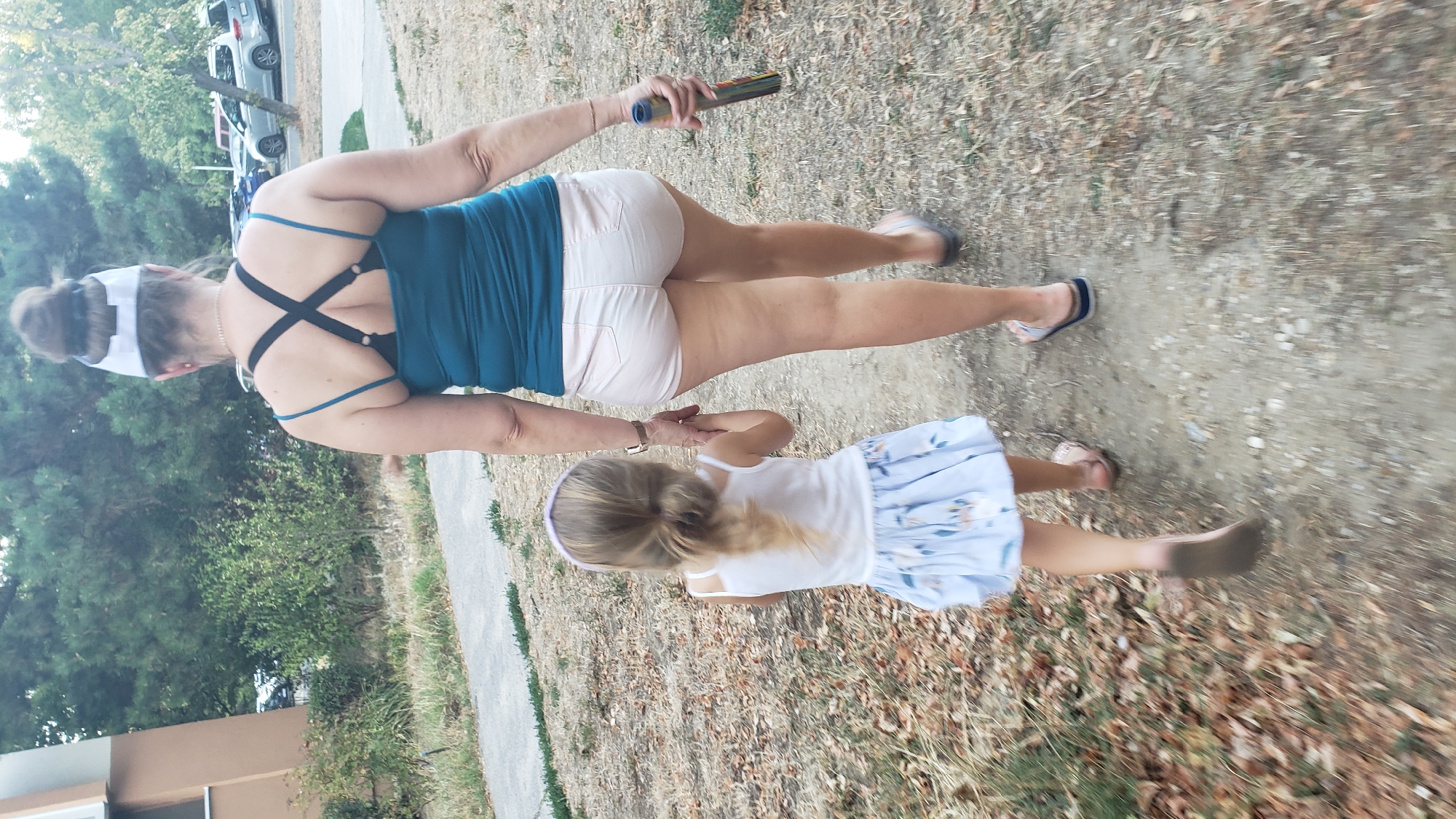 A woman and a young girl walk away from the camera on a dirt path lined with dry grass and leaves. The woman wears a teal tank top, pink shorts, and sandals. She holds a cylindrical object in her right hand. The girl wears a white sleeveless top and a light blue and white patterned skirt. She holds the woman's left hand and wears pink sandals. The background shows trees, a building, and parked cars, suggesting an outdoor urban environment. The lighting indicates it is daytime, likely late afternoon or early evening.