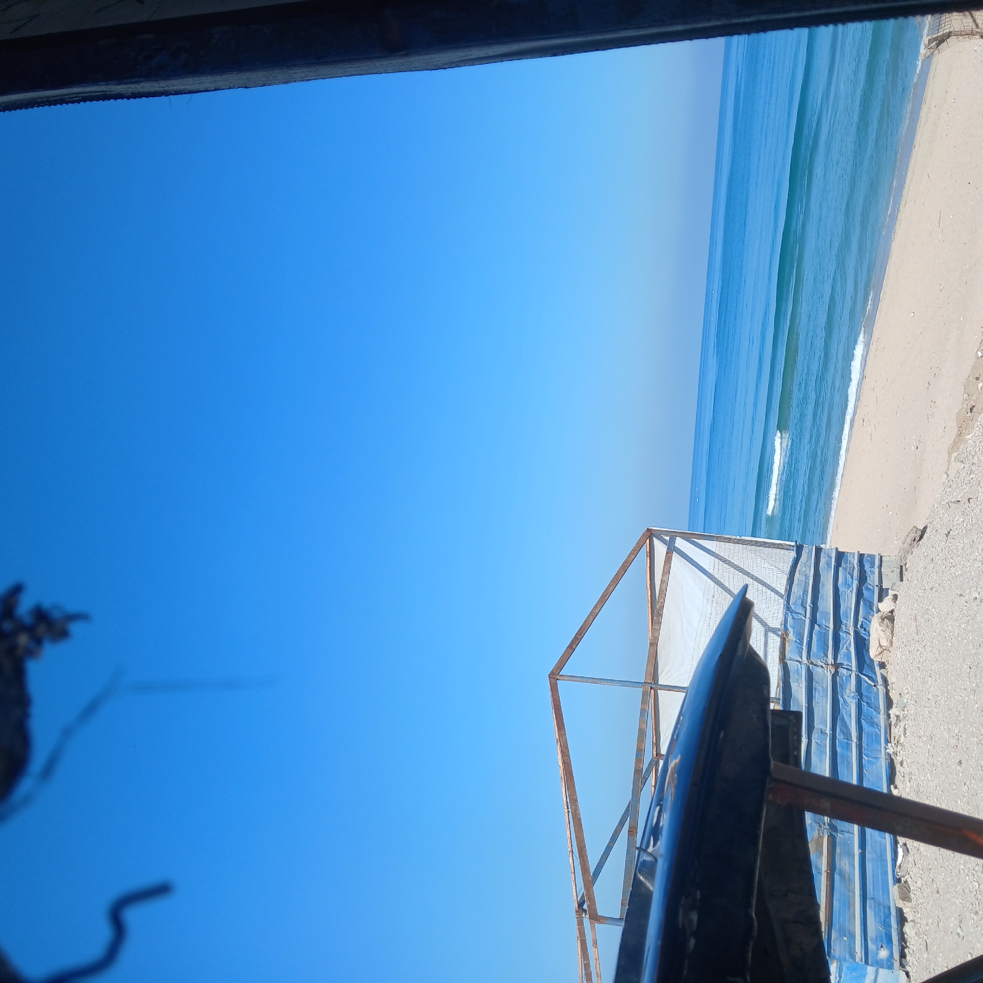The image depicts a bright, sunny coastal scene, viewed as if looking out from under a structure. The predominant feature is a vast, clear blue sky occupying the upper left portion of the frame. To the right, a blue-green ocean with visible waves meets a light-colored sandy or pebbly beach that stretches along the coastline.
In the immediate foreground, across the top edge of the image (after correcting for rotation), a dark, out-of-focus element, possibly an awning or a roof beam, frames the view. In the lower left foreground, another blurry, dark object, possibly foliage or a complex abstract shape, is visible.
A prominent structure made of rusty metal frames stands in the mid-ground, extending from the lower right towards the center. Part of this structure is covered with what appears to be blue corrugated metal or tarpaulin sheets. In the lower right foreground, a dark, curved object, possibly the bow of a small boat, a kayak, or a protective cover, is partially visible and in focus.
No people or explicit activities are taking place. The time of day appears to be mid-day, characterized by bright, clear sunlight and a cloudless sky, suggesting fair weather. There is no visible text in the image.