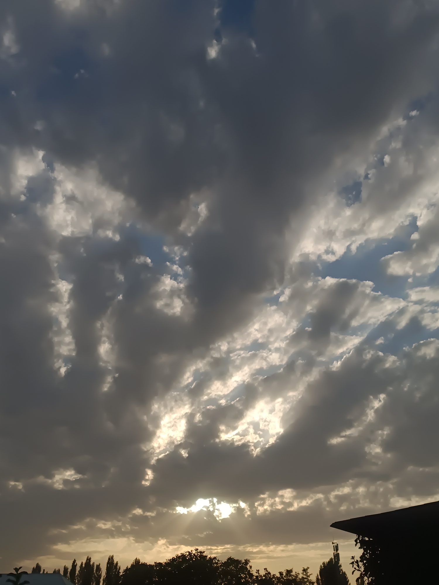 The image shows a sky filled with dramatic clouds, some dark and heavy, others lighter and reflecting sunlight. Sunlight is breaking through the clouds in several places, creating bright rays. In the lower portion of the image, a silhouetted roofline of a building is visible on the right, and a line of silhouetted trees and some smaller buildings are visible across the horizon. The overall impression is of a dynamic and somewhat overcast sky, possibly at dawn or dusk. There are no people or animals clearly visible. The scene appears to be an outdoor environment in a built-up area.
