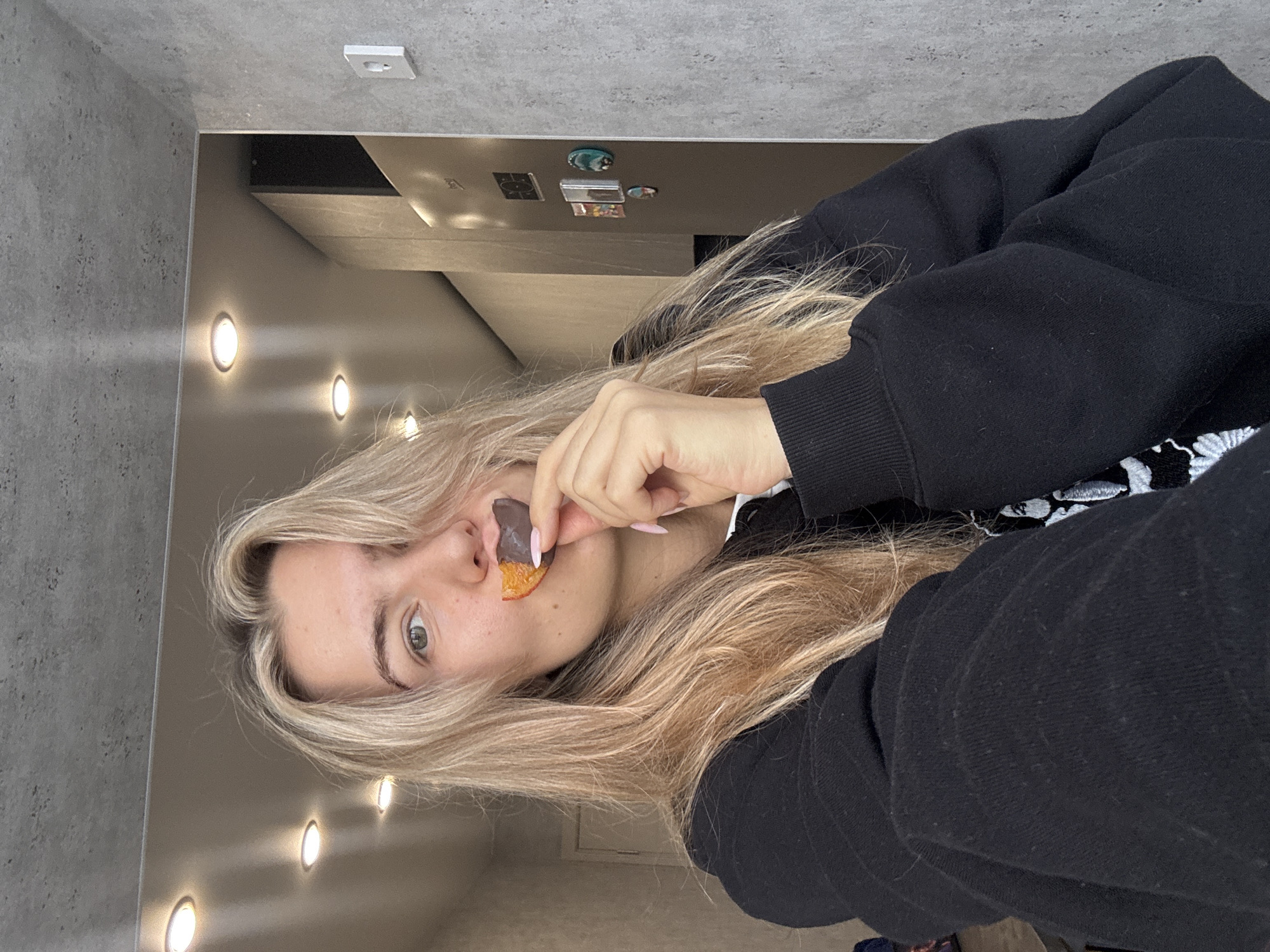 A young woman with long, straight blonde hair and fair skin with light freckles is depicted in an indoor setting. She has light-colored eyes and is looking directly at the camera with a neutral expression. She is wearing a black hoodie or sweatshirt, which covers her arms and torso. Her fingernails are painted a light, neutral pink.
In her right hand, she holds a piece of what appears to be dried orange fruit, possibly an apricot or mango slice, partially dipped in dark chocolate. She is bringing the snack towards her mouth.
The background features contemporary interior elements. To the left of the frame, a textured grey wall is visible. Behind the woman, there is a smooth, light-colored surface, possibly a cabinet or the side of a refrigerator, upon which several small, colorful magnetic items are affixed. Above this surface and behind the woman, several circular recessed lights are embedded in a light-toned ceiling or upper wall, casting bright, warm light. An electrical outlet or light switch is mounted on a grey wall section further back. The overall lighting suggests an indoor setting, likely a modern kitchen or living area. There is no visible text in the image.