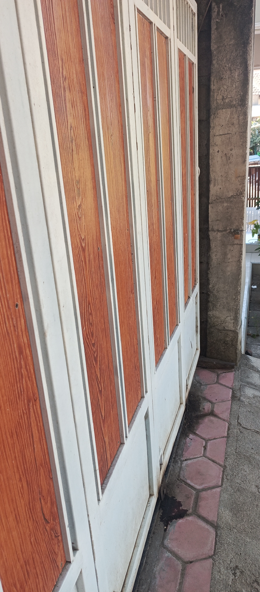 This is a low-angle, full shot of a white metal gate with wooden-patterned panels, set against a concrete pillar. The gate is positioned in Arjasa, Indonesia. The wooden panels are a warm, reddish-brown with a visible wood grain texture. The gate is composed of vertical panels, alternating between the wooden and white metal sections, with thin white vertical bars at the top. The ground in front of the gate is a combination of grey concrete and hexagonal pink pavers. There are dark patches on the ground near the base of the gate, possibly indicating water or dirt. In the background, to the right of the gate, a section of a building and some greenery are visible. The lighting suggests it is daytime, with shadows cast by the gate. No people are present in the image.