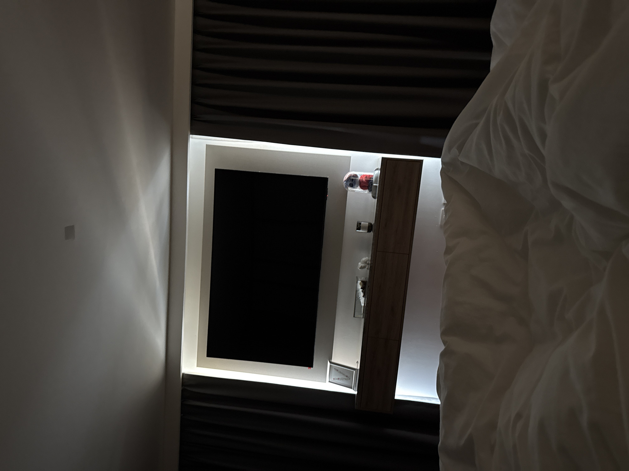 The image depicts a dimly lit interior with a focus on a wall-mounted television. The TV screen is black, suggesting it is either off or displaying a dark image. Adjacent to the TV is a vertical wooden panel with several small objects mounted on it, potentially decorative items or control devices. A section of what appears to be a bed with white bedding occupies the right side of the frame. The wall surrounding the TV and panel is off-white and illuminated by a soft, indirect light source, creating a halo effect around the recessed display. The environment suggests a modern hotel room or bedroom setting. The image was not taken in San Bernardino Tlaxcalancingo, Mexico.