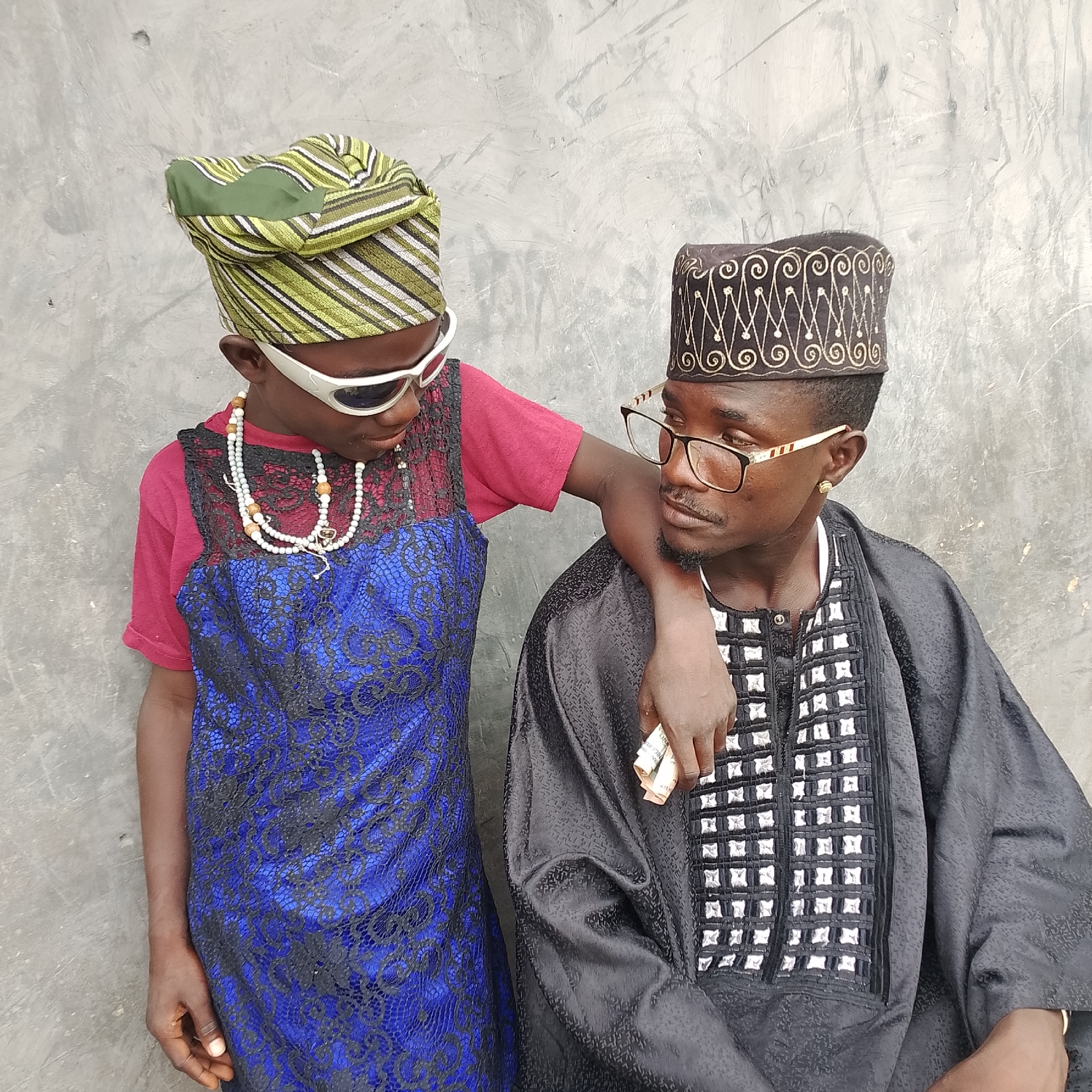 Two individuals stand in front of a textured gray wall. The person on the left, appearing younger and wearing a green and white striped headwrap, white sunglasses, and a blue and black lace dress over a red shirt, leans their head towards the person on the right. They also wear a multi-strand white beaded necklace. The person on the right, wearing a dark brown cap with gold embroidery, glasses with gold accents, and a black embroidered garment, is looking towards the left person. The right individual's right arm rests on the left individual's shoulder, and they are holding a wad of Nigerian currency in their right hand. The wall behind them has subtle textures and some faint markings.