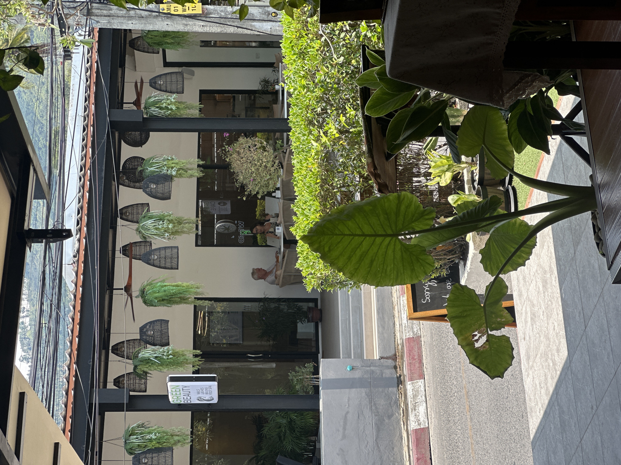 The image depicts an outdoor scene in Baan Chaweng Noi, Thailand, presenting a partial view of a commercial establishment identified as "GREEN BEAUTY," offering services such as nails, lashes, waxing, and foot spa. A sign with the text "Sorry, 1.6 are MOED CVLE" is visible.
The building features a modern design with a black metal framework, visible hanging plant decorations, and open-front facade. Two people are visible inside the establishment.
The surrounding environment includes foliage, such as large-leafed plants in the foreground and a hedge along the side. The presence of a road with red and white lane markings is also noted, along with a street sign.
