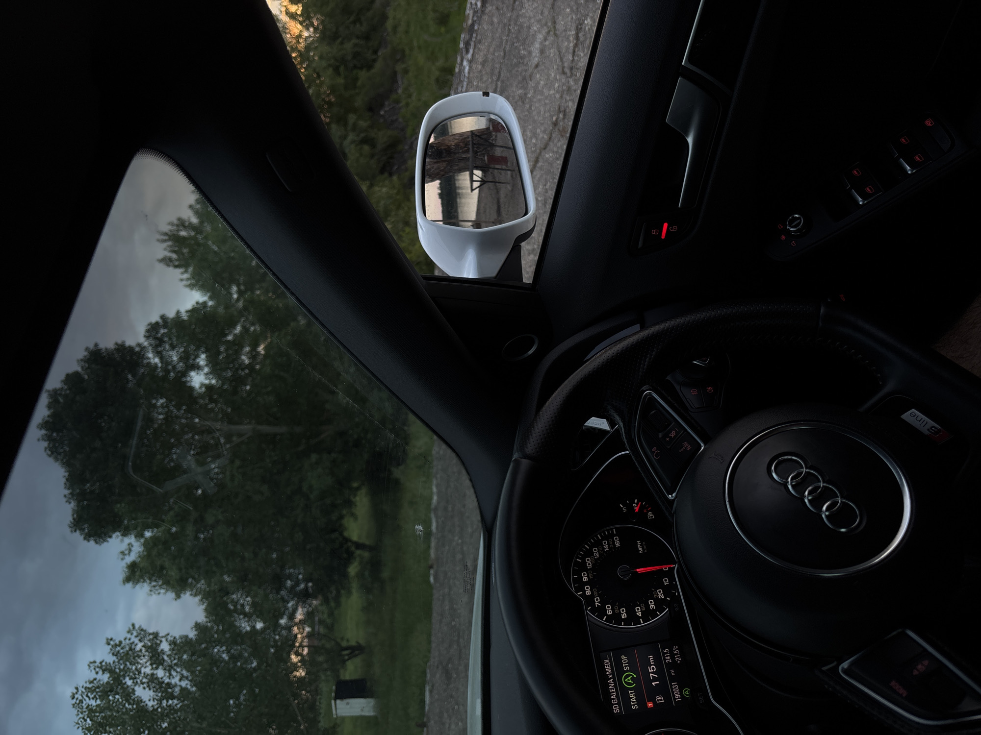 The image depicts the interior of an Audi car. The dashboard and steering wheel are prominent, with an Audi emblem visible on the steering wheel. The instrument panel indicates a speed of around 10 mph, with a mileage of 175 miles and an outside temperature of 21.5°C. Through the front windshield, trees are visible. The driver's side mirror is in view, reflecting an external scene. The location in the city of Dolni Tsibar, Bulgaria cannot be confirmed solely based on the image.