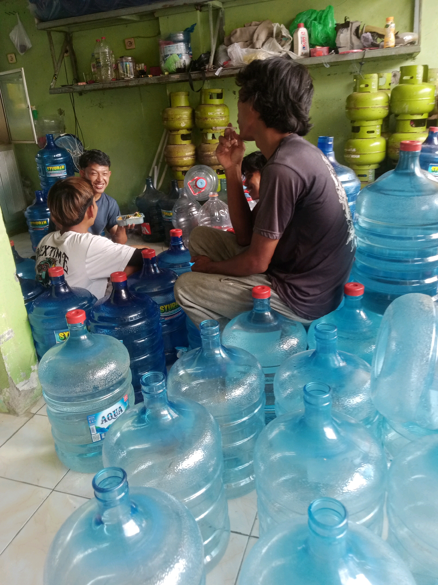 The image depicts a storage area filled with numerous large, clear blue plastic water jugs, commonly used for dispensing purified water. Several of these jugs are full and stacked, while others appear empty or partially filled. The environment is cluttered, with shelves in the background holding various items, including plastic bottles, what appear to be cleaning supplies, and possibly food items. Stacked in the background are several green propane gas cylinders.
In the foreground, three individuals are present. Two young men are seated on the floor, engaged in conversation. One of them is holding a small tray with what seems to be food. A third individual, also seated, is positioned to the right, facing away from the camera and towards the water jugs. He appears to be smoking. The floor is tiled and slightly wet in some areas. The walls are painted a light green color. The scene suggests a small business or warehouse operation likely related to water distribution, possibly located in Grendeng, Indonesia, as indicated by the presence of propane cylinders often used in such regions for various purposes, including cooking and potentially business operations. The overall atmosphere is casual and functional.