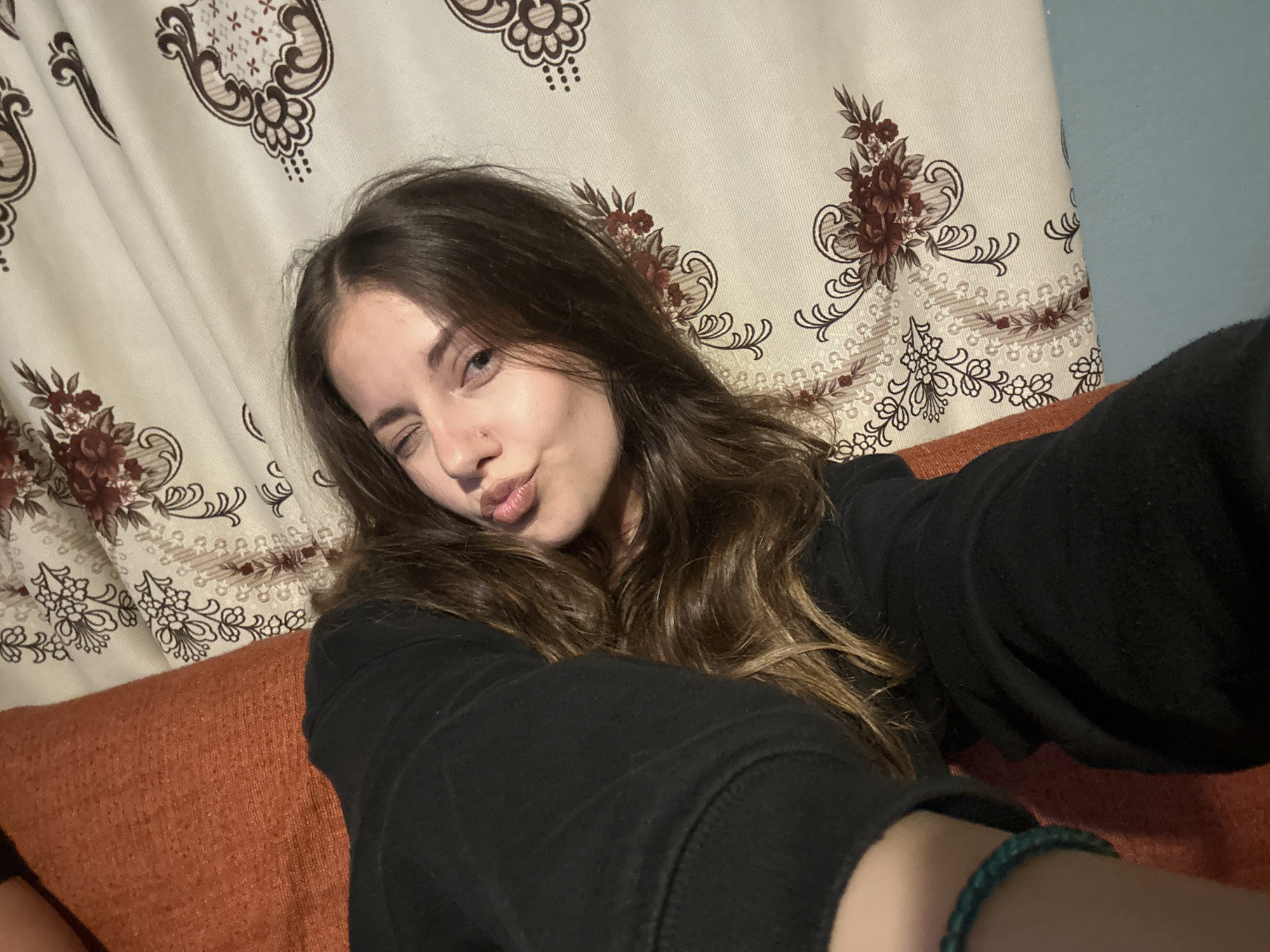 A young female with long brown hair poses for a selfie, partially cropped at the torso. She wears a black long-sleeved top and a turquoise braided bracelet on her right wrist. Her right eye is open and directed towards the camera, while her left eye is winked shut, and her lips are pursed in a pouting expression. A small metallic stud piercing is visible on her left nostril. She is seated on an orange, textured fabric couch or cushion, visible in the lower foreground and midground. The background consists primarily of a cream-colored curtain adorned with a repeating pattern of stylized floral and paisley motifs in dark brown and maroon. These motifs include large flower clusters, scrollwork, and small 'X' shapes within some elements. A narrow vertical section of a light blue wall is visible to the far right of the curtain. The indoor setting is in the city of Unknown, Unknown.