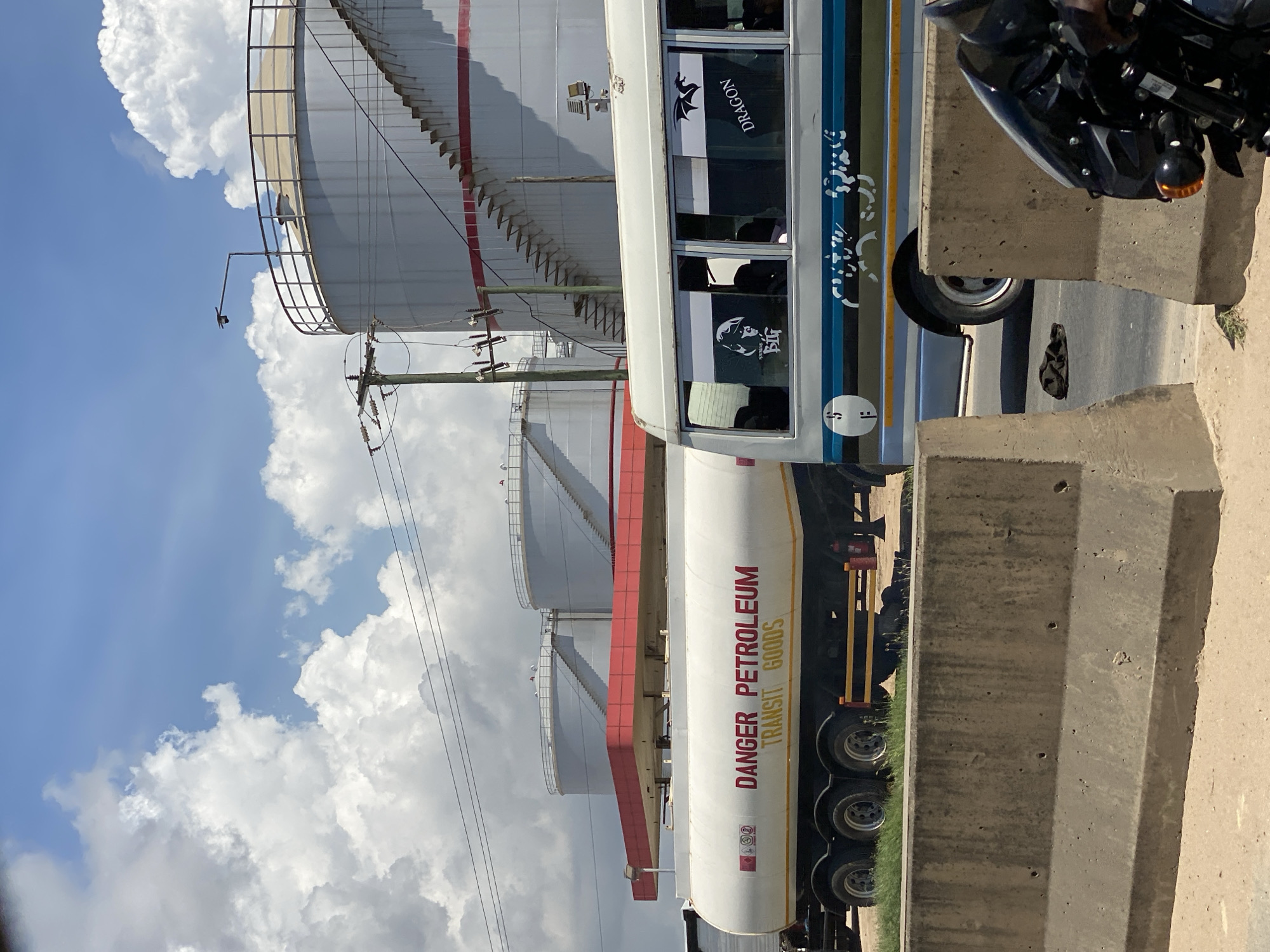A tanker truck, labeled "DANGER PETROLEUM," is parked alongside what appears to be a road divider. The surroundings suggest an industrial area, featuring large, cylindrical storage tanks with exterior staircases. The sky above has a mix of blue and white clouds. The location is in Dar es-Salaam, Tanzania, based on visual cues. A passenger bus is next to the tanker truck and a motorcycle is parked next to a curb.