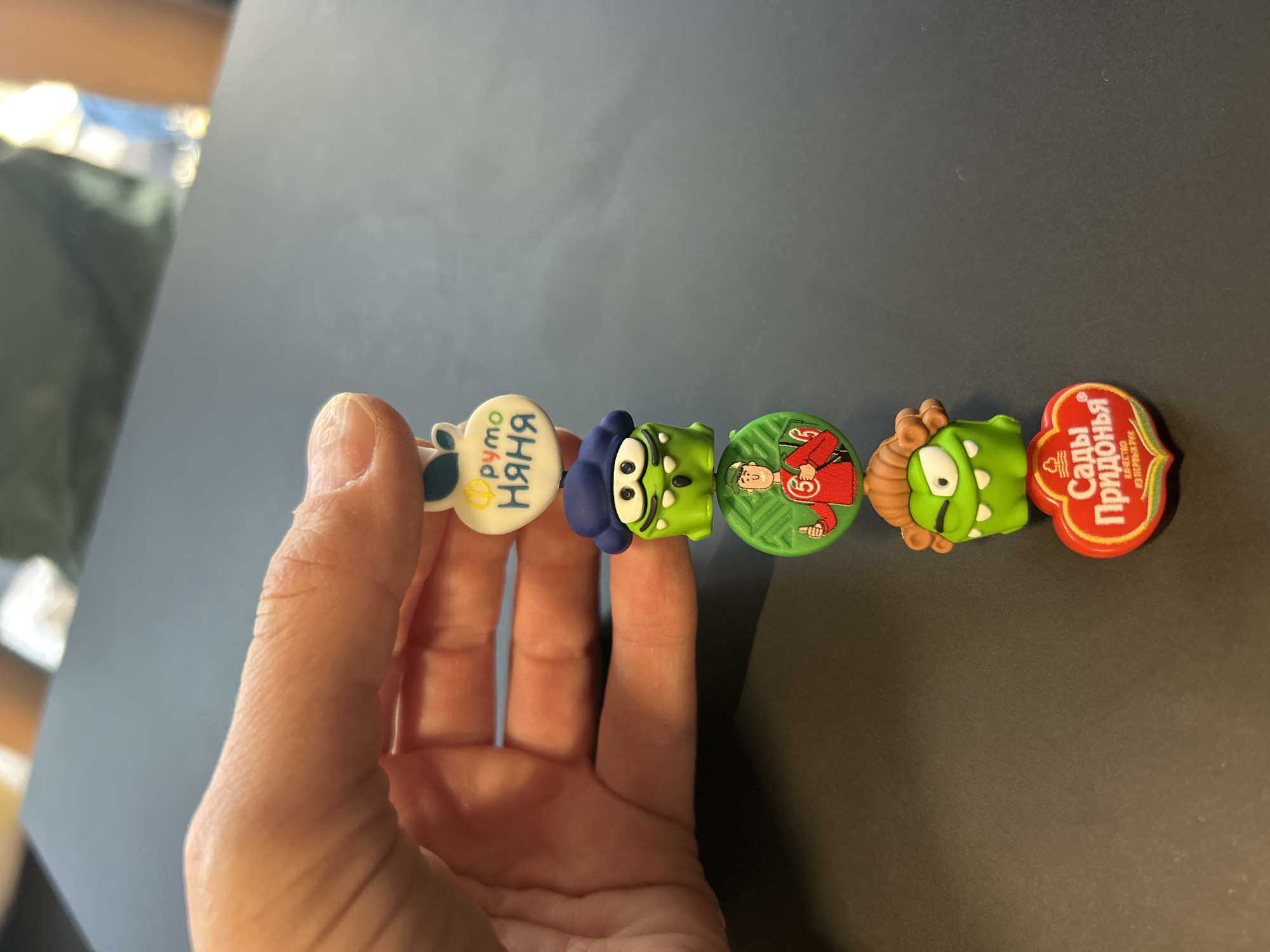 A person holds a row of five novelty refrigerator magnets, with a black surface visible in the background. From left to right, the magnets are: a white cloud with the inscription "Pimo Nyanya"; a green monster with a blue hat; a green circular magnet depicting a person in a red and green outfit with the number 5; a green monster with brown hair; and a red magnet with the inscription "Sady Pridonya". The location is Izhevsk, Russia, based on the Russian text on the magnets.