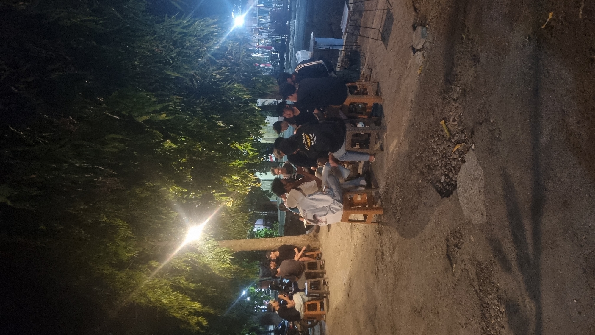 The image depicts an outdoor gathering of young men at night in Kepanjen, Indonesia, likely at a casual eatery or roadside stall. Several groups of individuals are seated around tables, engaged in conversation. The setting is illuminated by overhead lights, casting a warm glow and creating noticeable lens flares. Vegetation, including a large tree, is visible overhead and behind the people. The ground appears to be a mix of dirt and asphalt with some visible patches of damage or unevenness. In the background, there are indications of structures and possibly more tables and chairs, suggesting a public space where people congregate. The individuals are casually dressed, and their interactions appear relaxed and social.