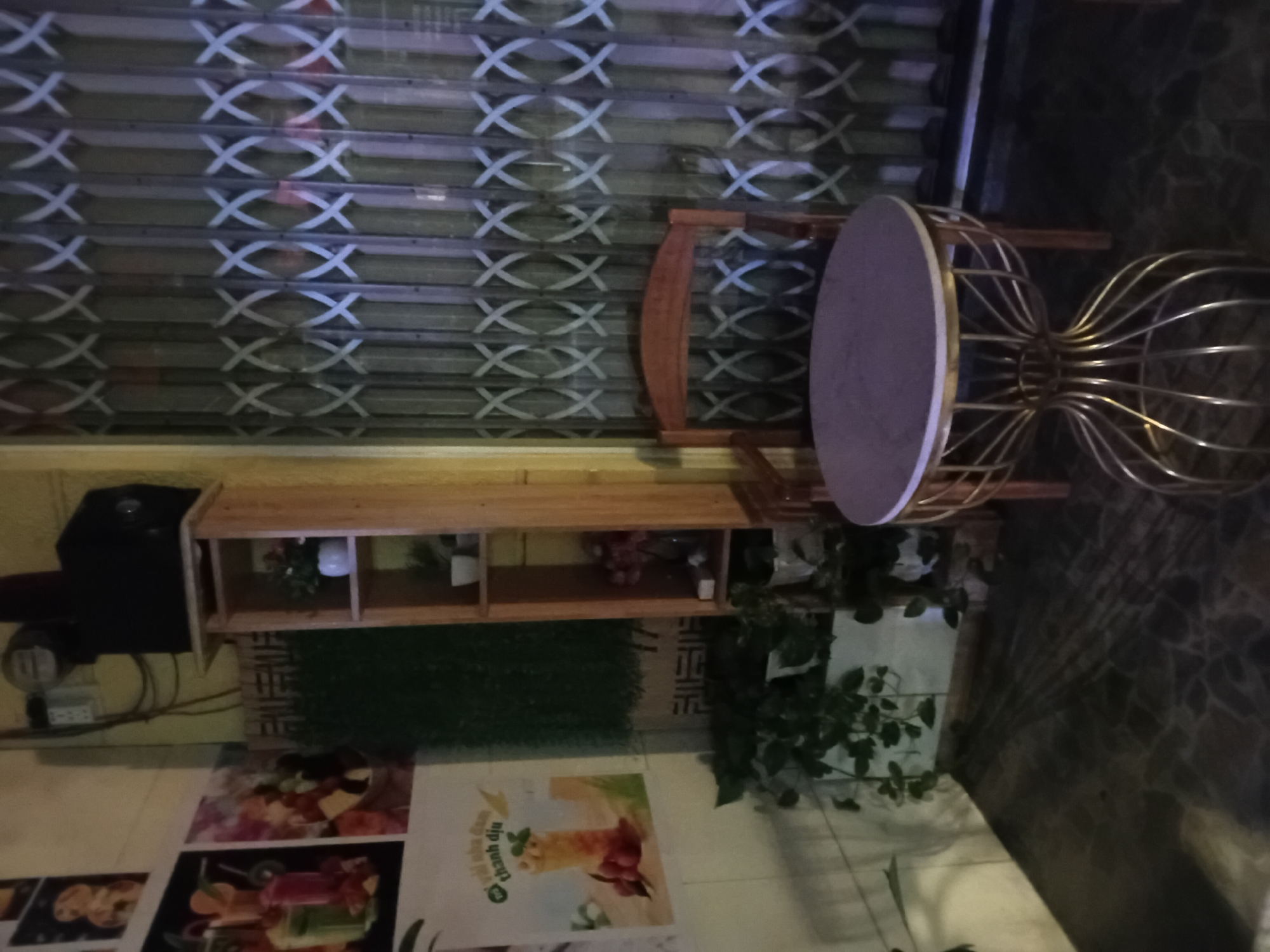 The image shows an indoor scene, likely a cafe or a shop, with a metal roll-up shutter in the background. In the foreground, there is a wooden shelf unit containing various items, including what appears to be decorative plants and possibly some beverages. To the right of the shelf, there is a round table with a light-colored top and a distinctive gold wire base, accompanied by a wooden chair. In front of the shelf, a piece of artificial turf is visible. On the floor, several posters are displayed, some featuring images of food and drinks. One poster has Vietnamese text, including the words "thanh diệu". The overall lighting suggests it might be evening or an indoor setting with artificial lights. The environment appears to be clean and organized, with hints of greenery from the plants. The presence of posters and a table with chairs suggests a commercial establishment.