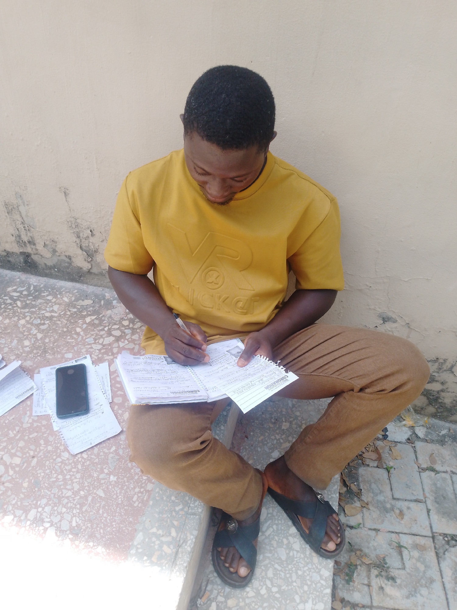 A young Black man wearing a yellow t-shirt and brown pants is sitting on a concrete ledge against a cream-colored wall, writing in a notebook. His head is bowed as he focuses on his task, a pen in his right hand. Next to him, on the ground, is a smartphone and several loose papers. The ground beneath him is a speckled concrete surface with a few fallen leaves scattered about.